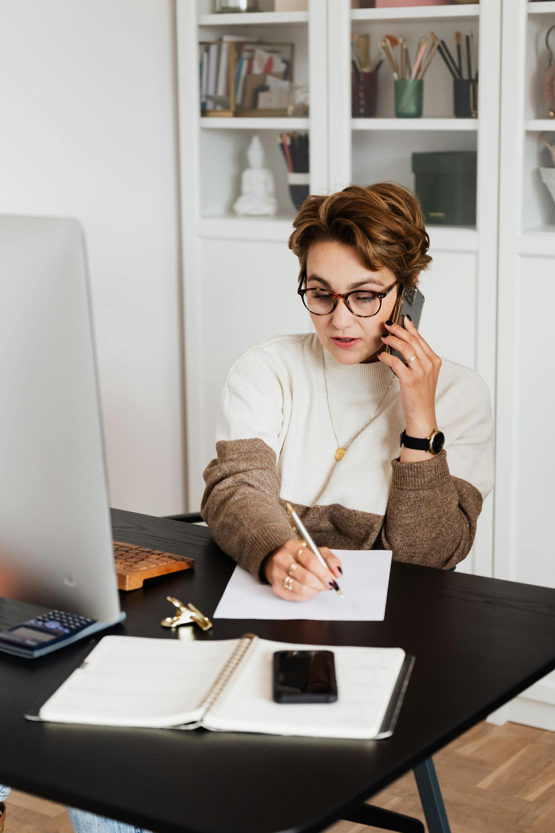 Person writing at a desk while on the phone in a bright home office