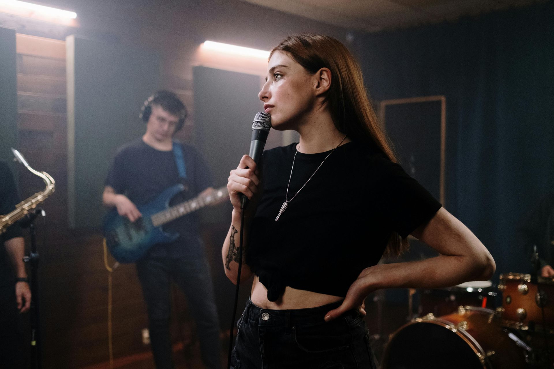 Singer holding a microphone in a dim rehearsal studio with a guitarist and drum kit behind her