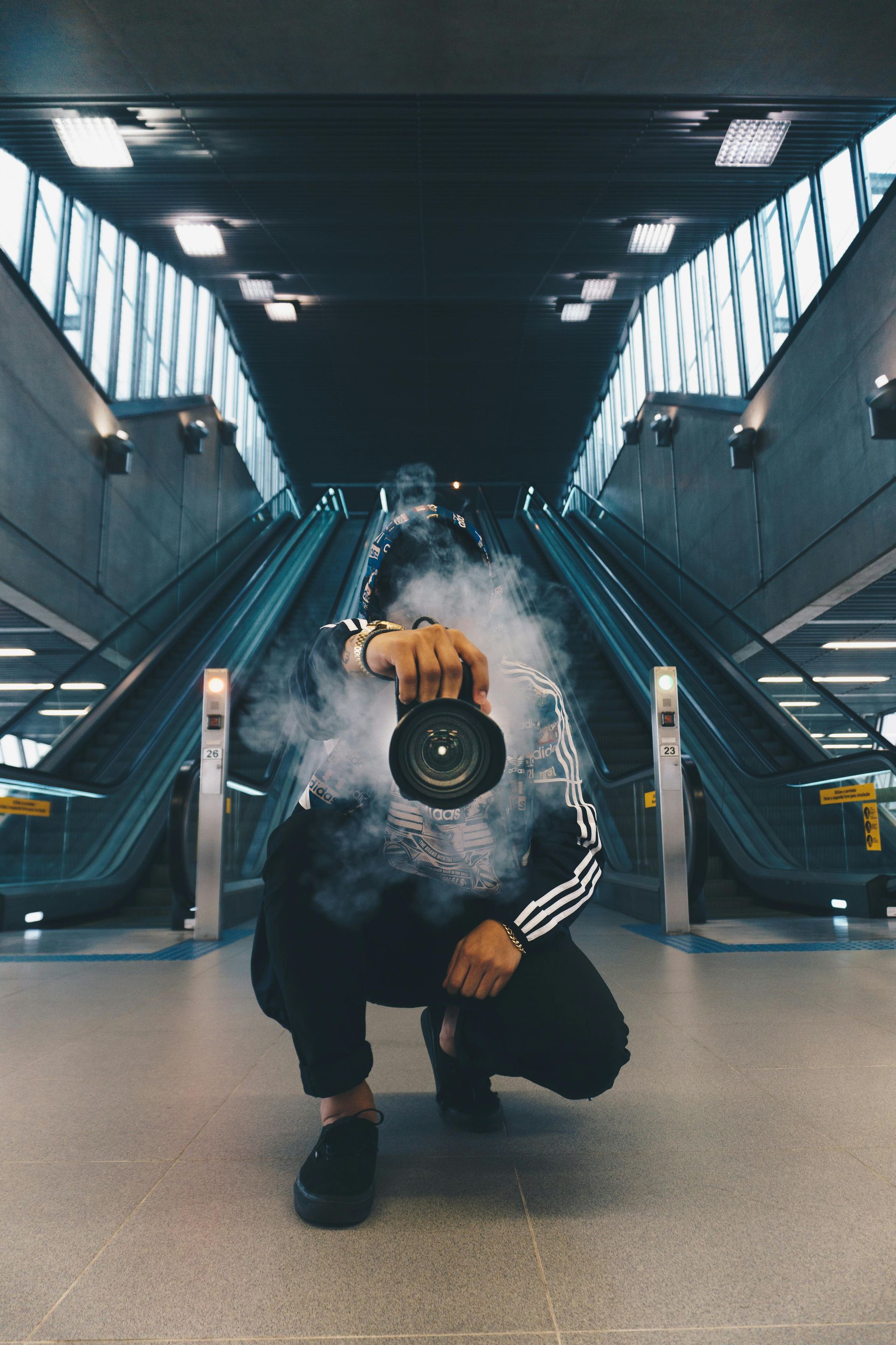 Person crouching in a modern train station, holding a camera, with escalators and skylights behind.