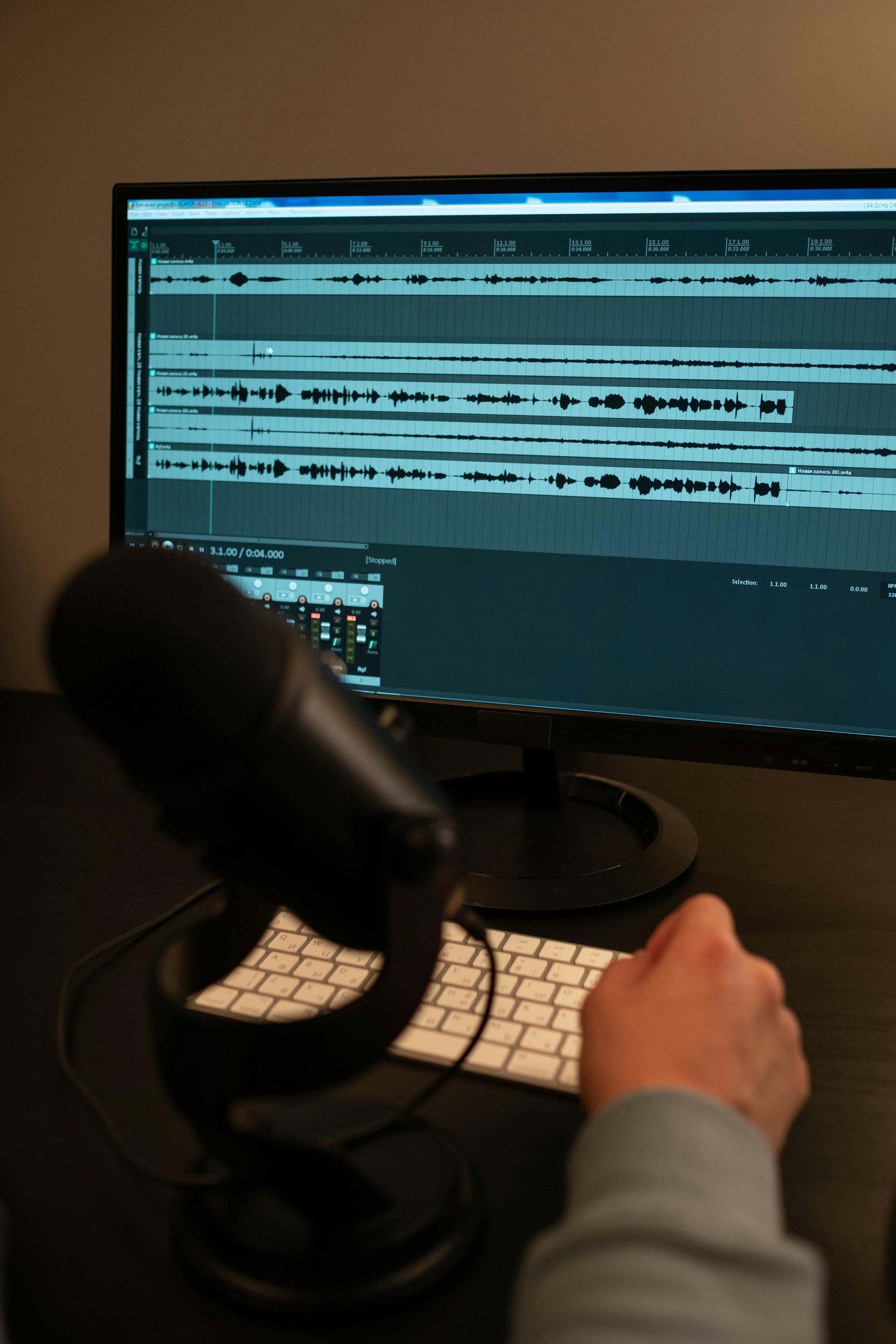 Person editing audio on a computer with waveform tracks, a microphone in the foreground.
