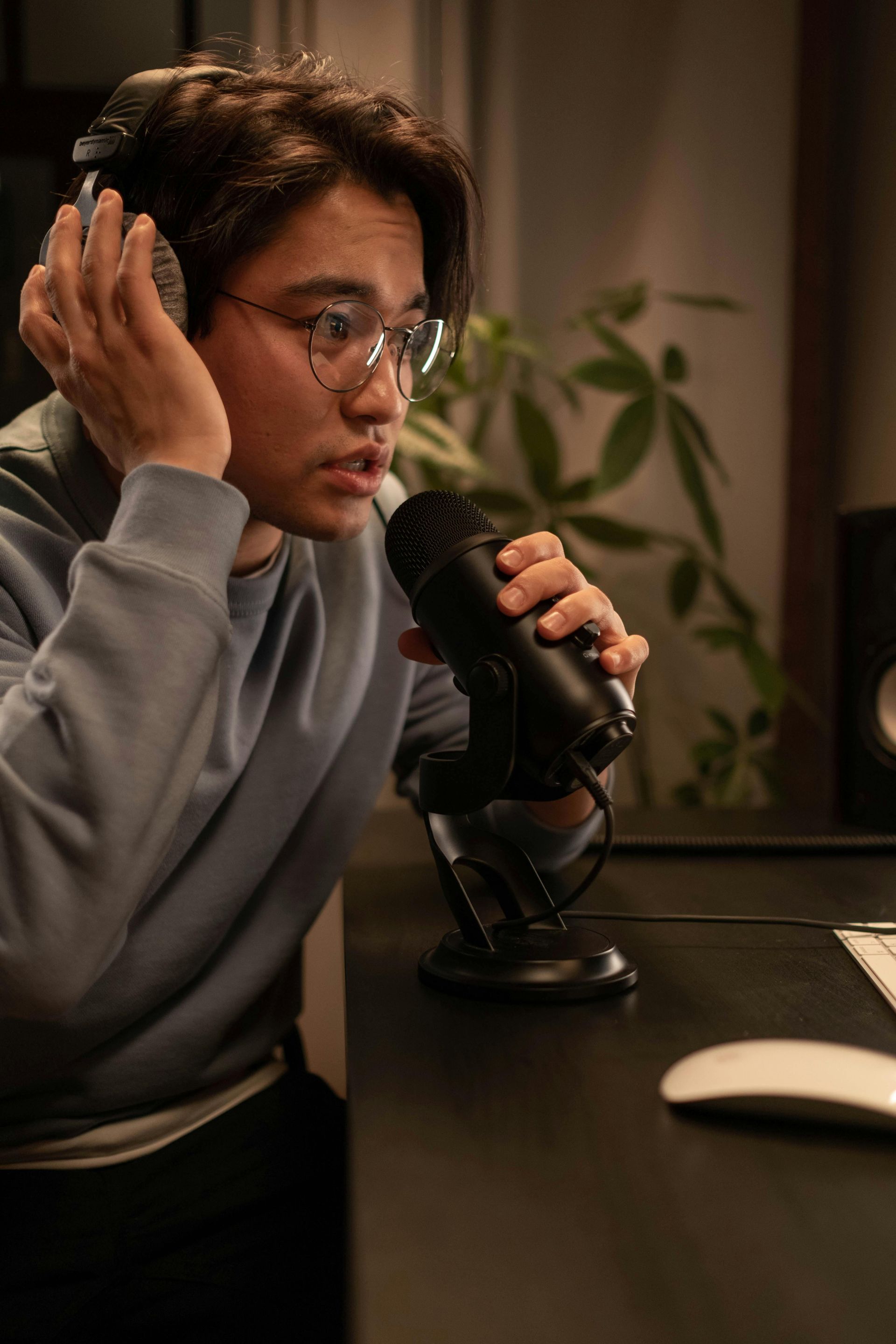 Person in glasses speaking into a microphone at a desk in a softly lit room