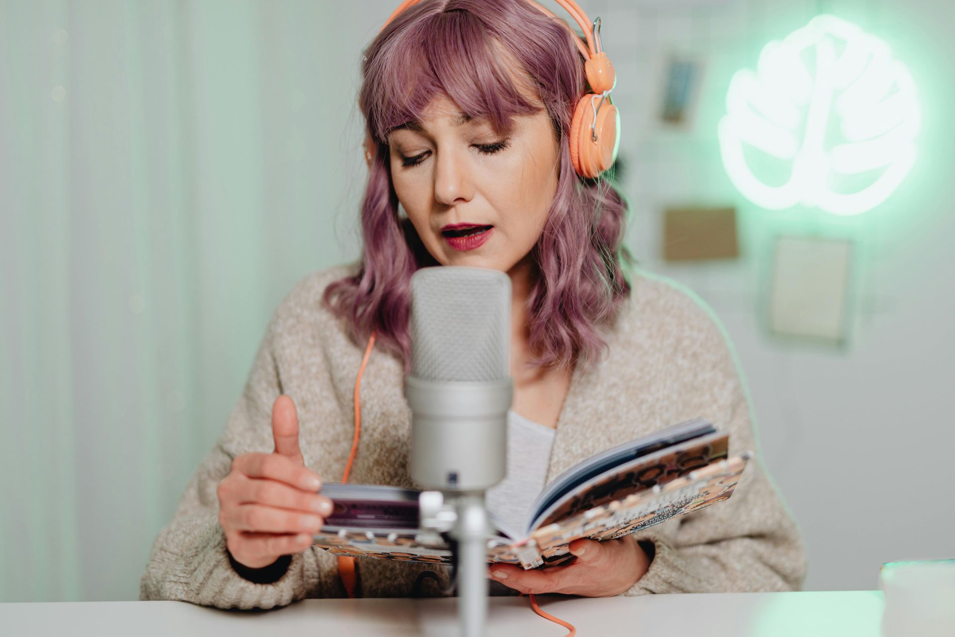 Woman with pink hair wearing headphones, reading aloud into a microphone at a desk in a soft-lit studio.