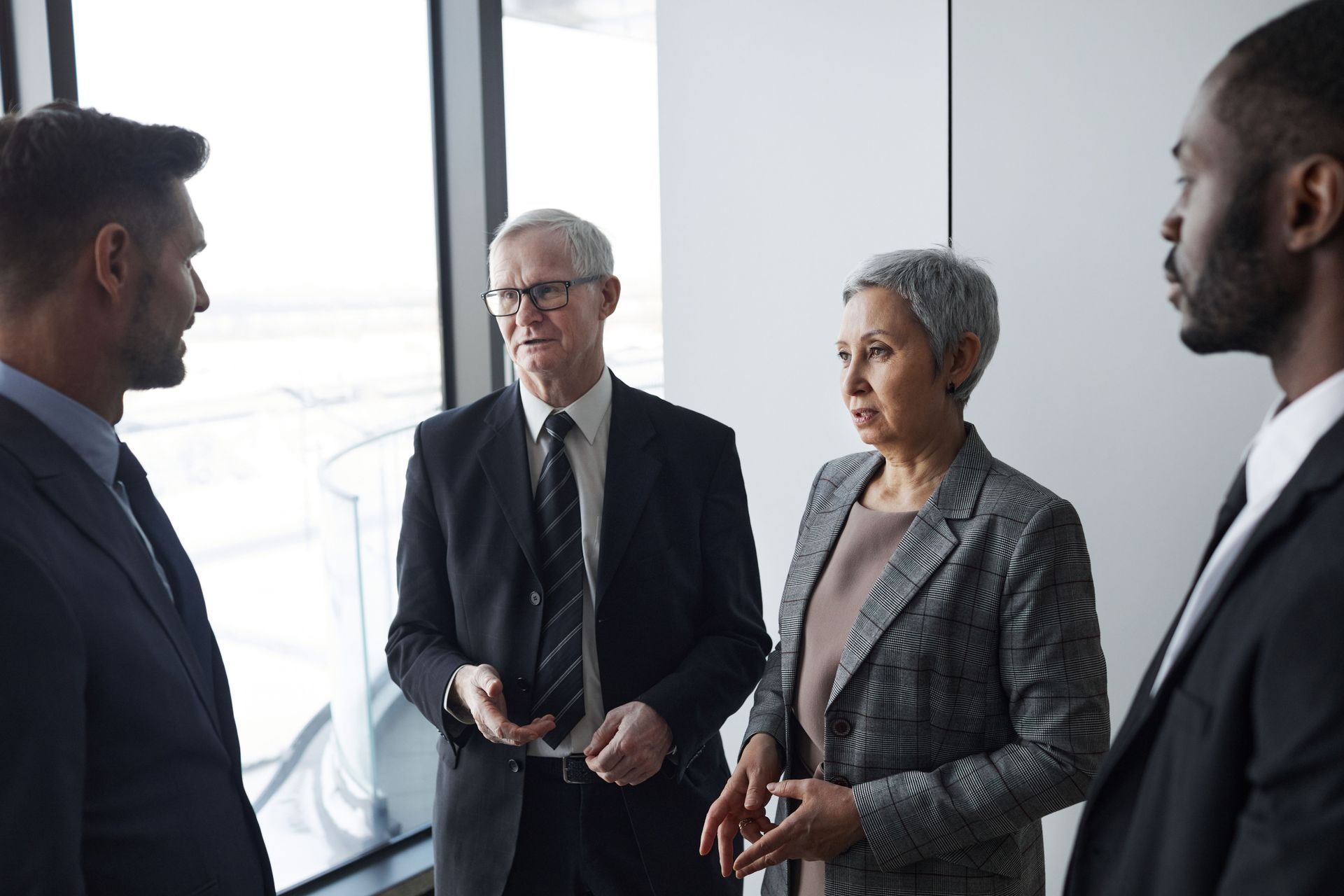 Four business professionals in suits talking in a bright office by a window.