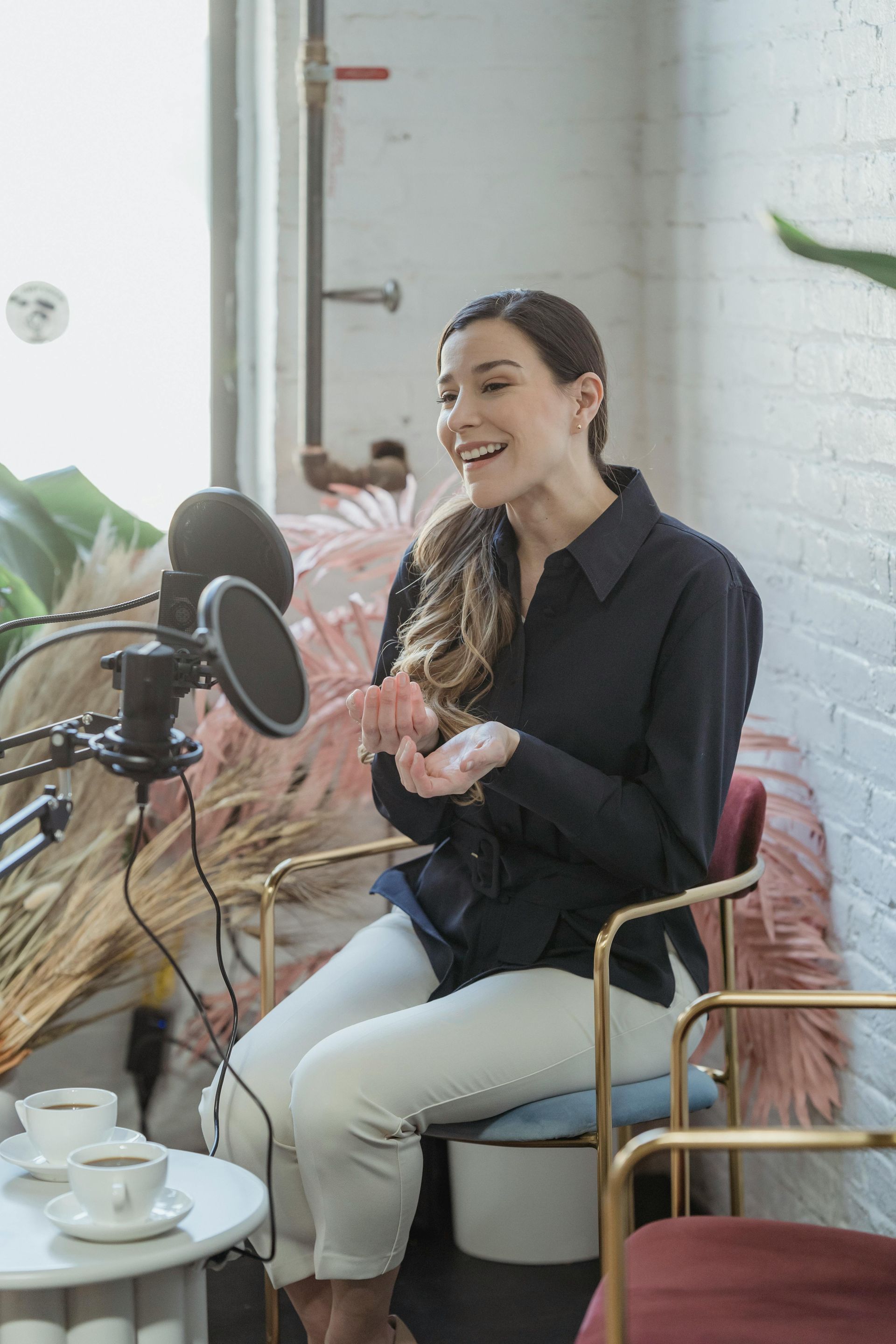 Woman speaking into a microphone in a bright café, seated by a table with a laptop and coffee cup.