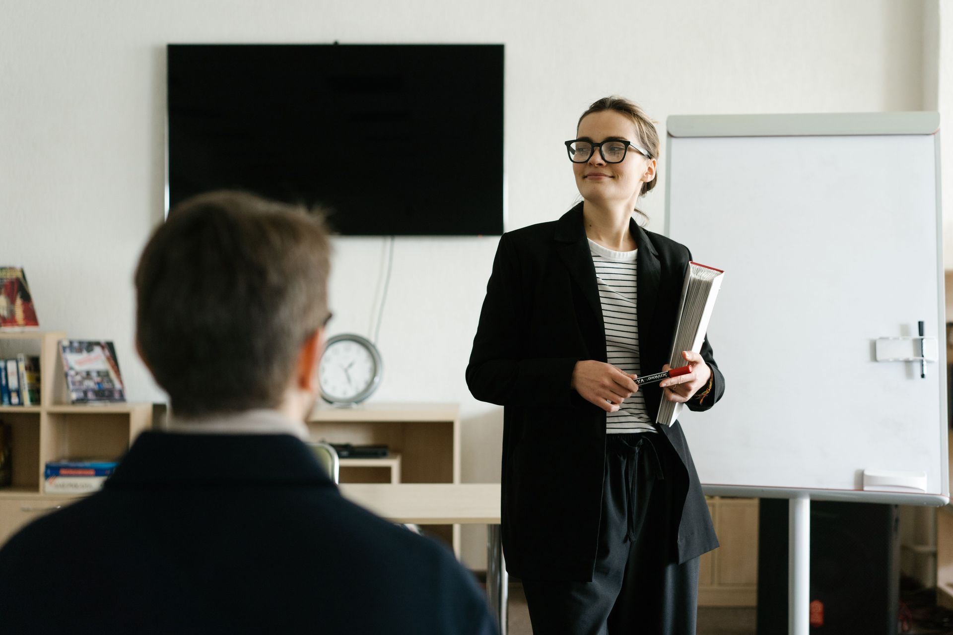 Presenter speaking in a meeting room beside a whiteboard, with a seated audience member in the foreground.