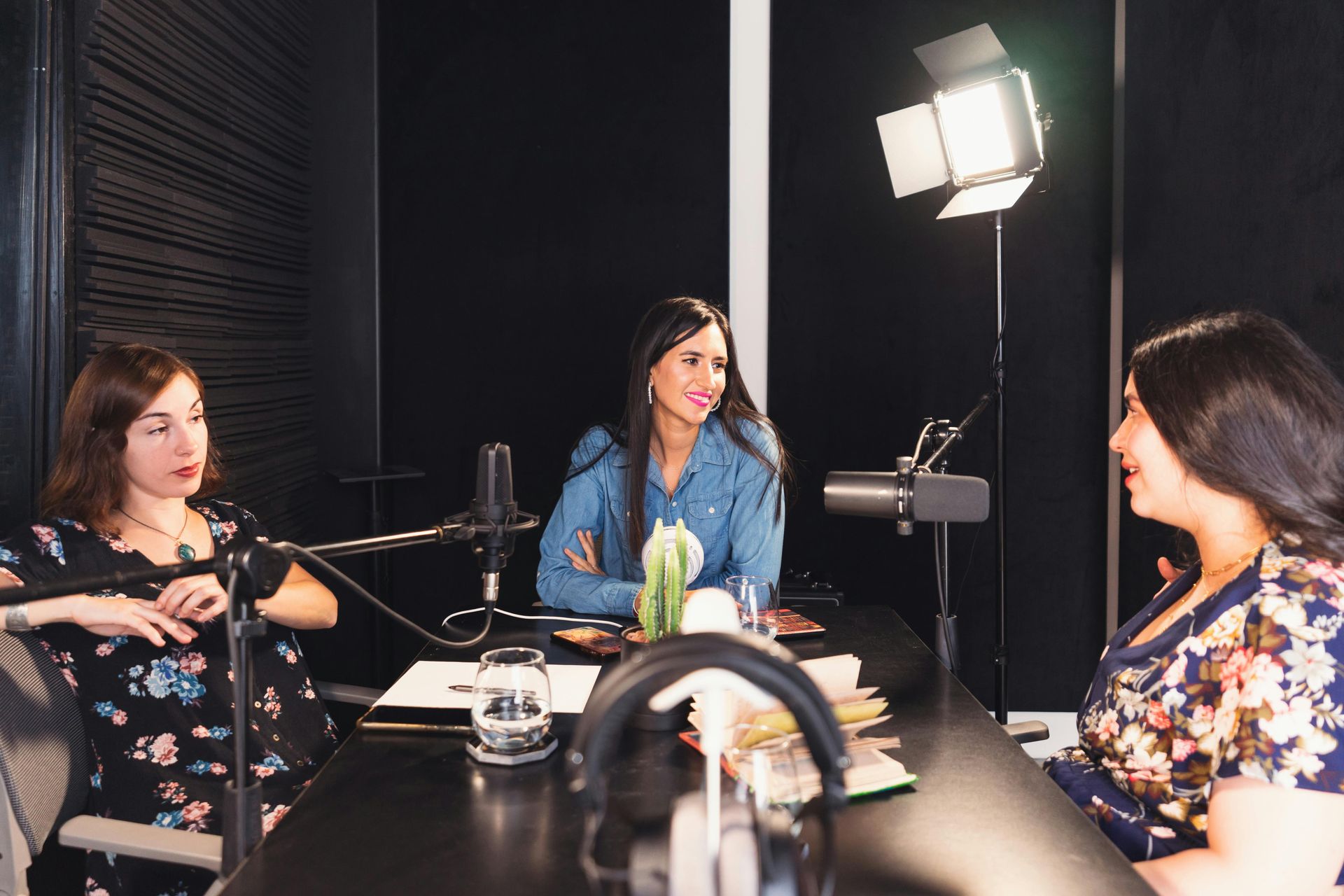 Three people in a podcast studio seated around microphones and a table under bright studio lights