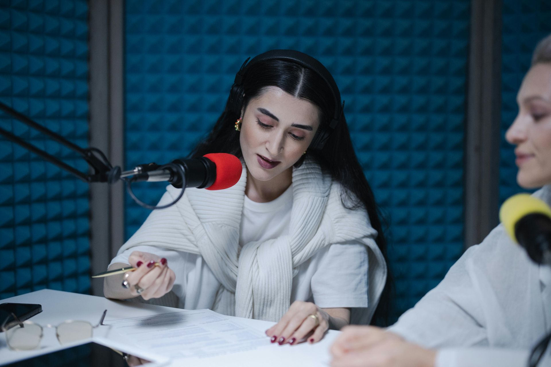 Two women recording a podcast in a studio, speaking into colorful microphones over papers