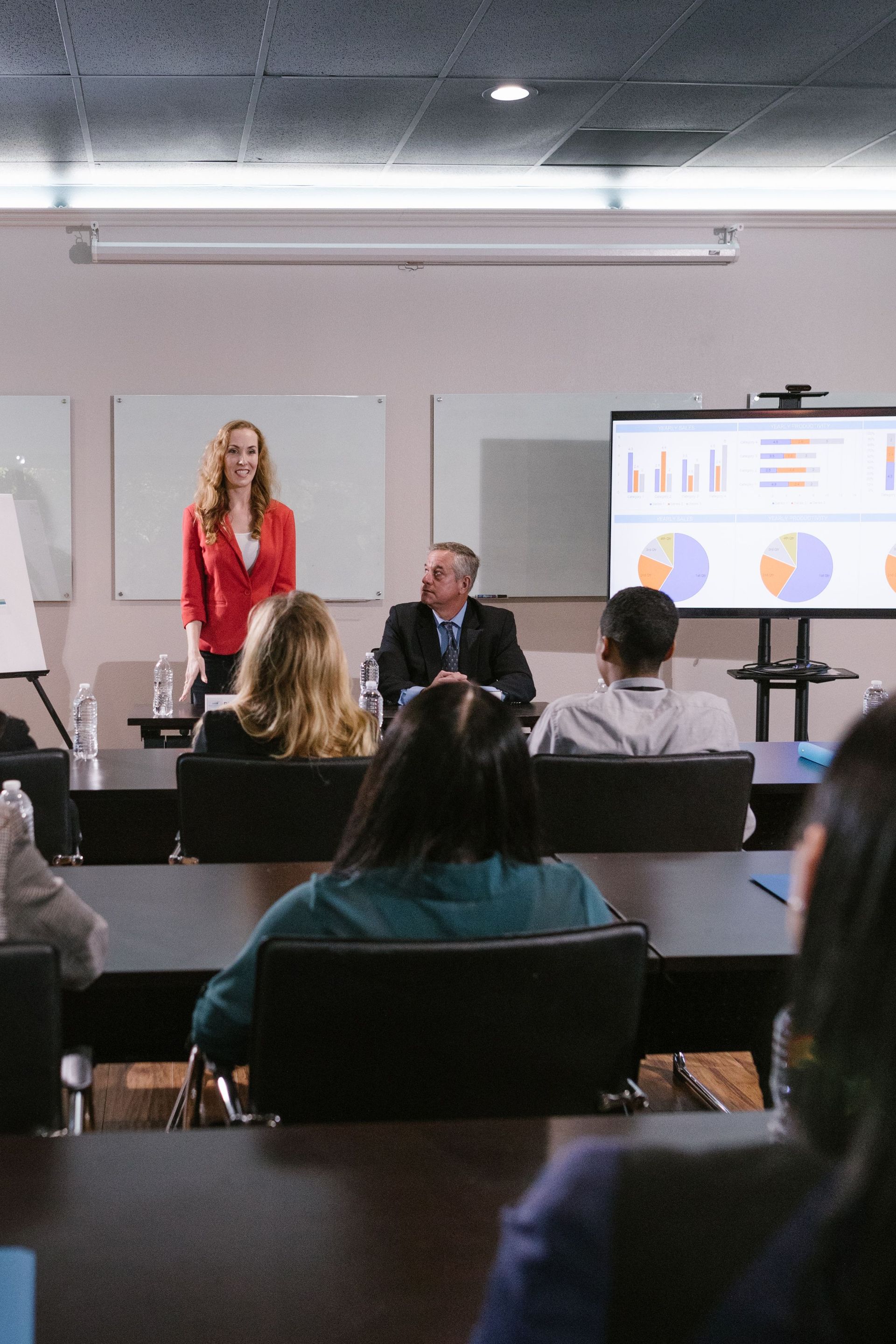 Presenter speaking in a meeting room while a slide deck is projected and attendees watch from rows of seats