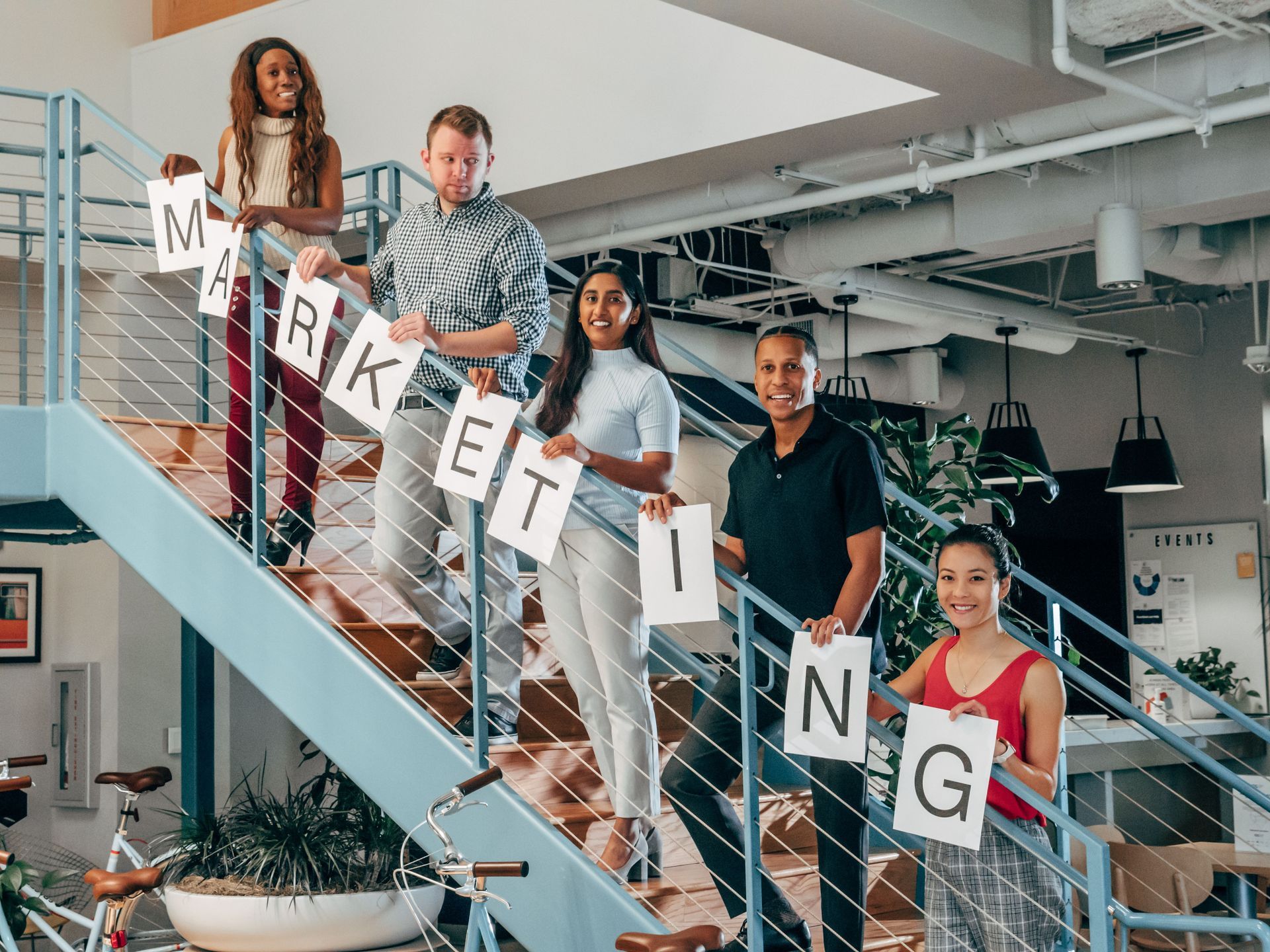 Four coworkers on stairs holding letter cards that spell “MARKETING” in a bright office.