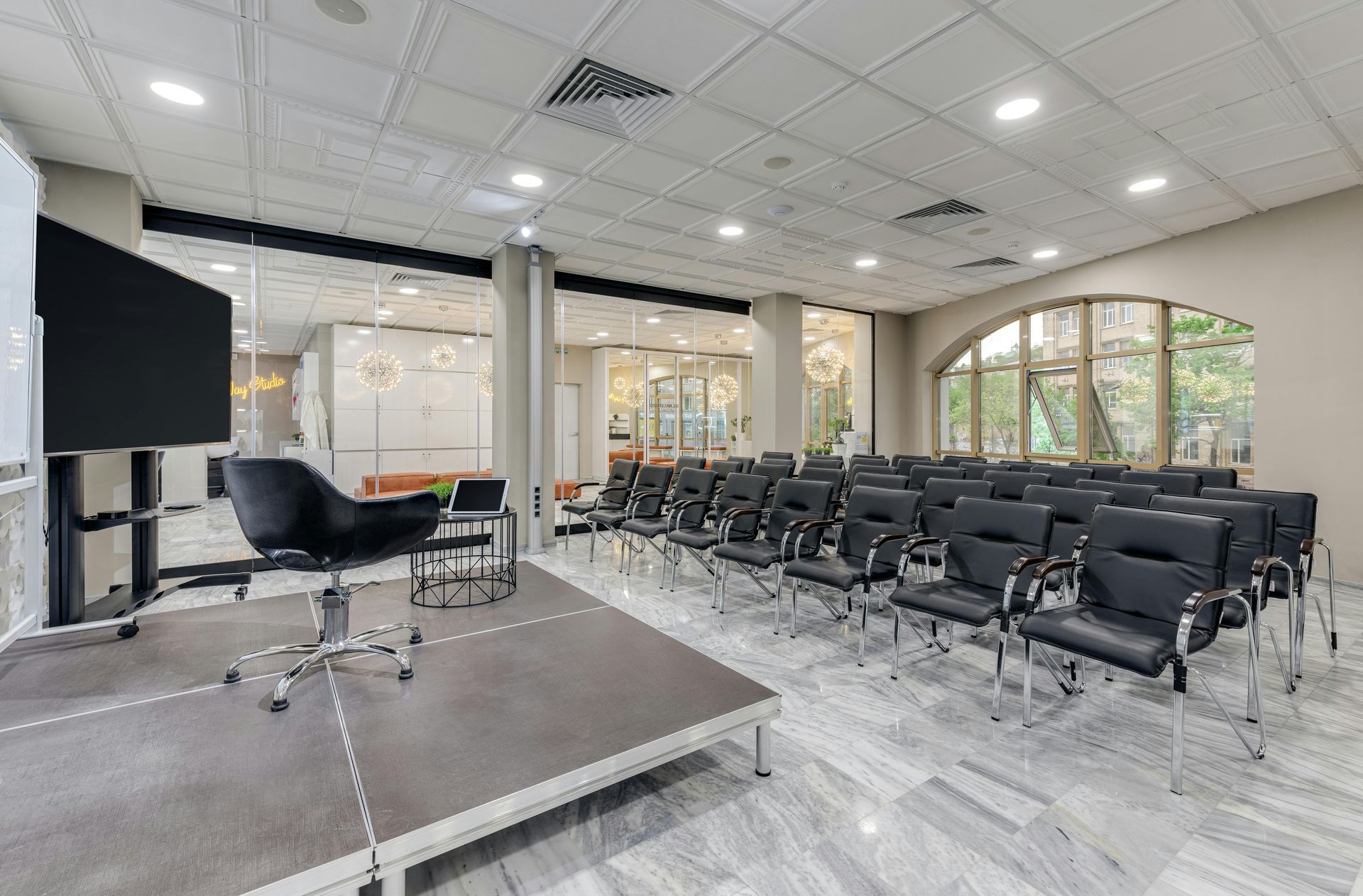 Empty conference room with rows of black chairs, a stage, and large windows under bright ceiling lights
