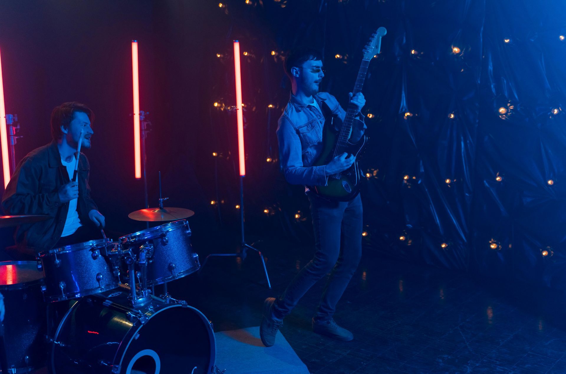 Two musicians perform on a dark stage with blue lighting and neon red lights