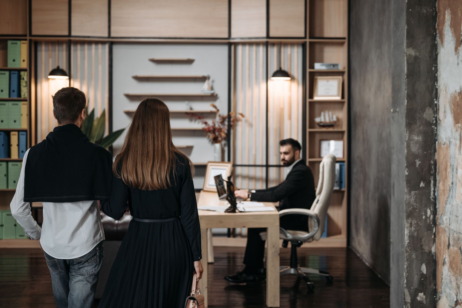 Two people entering a modern office reception area with a seated man at a desk and hanging pendant lights
