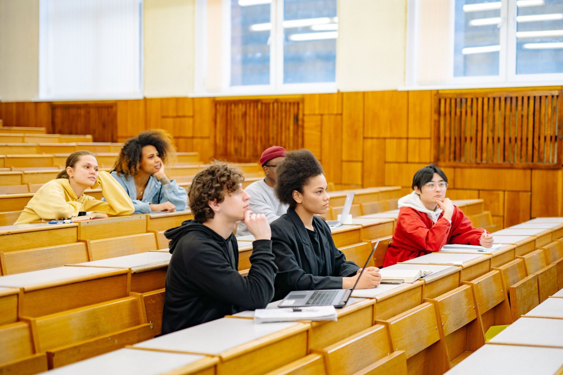 Students seated in a lecture hall, writing and listening at wooden desks.