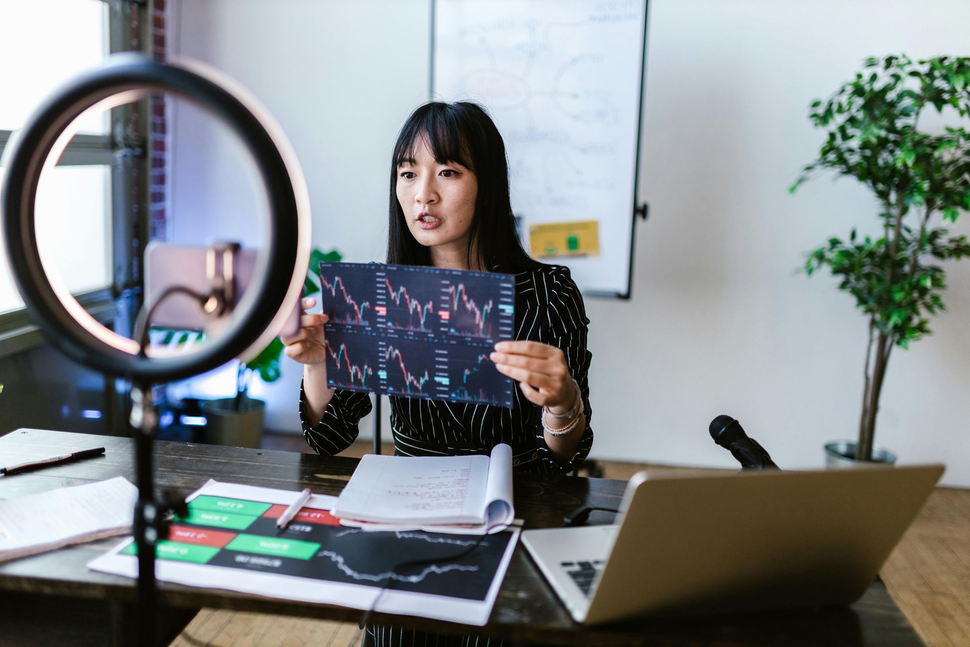 Person holding a tablet with a financial chart, seated at a desk in an office with a laptop and ring light.