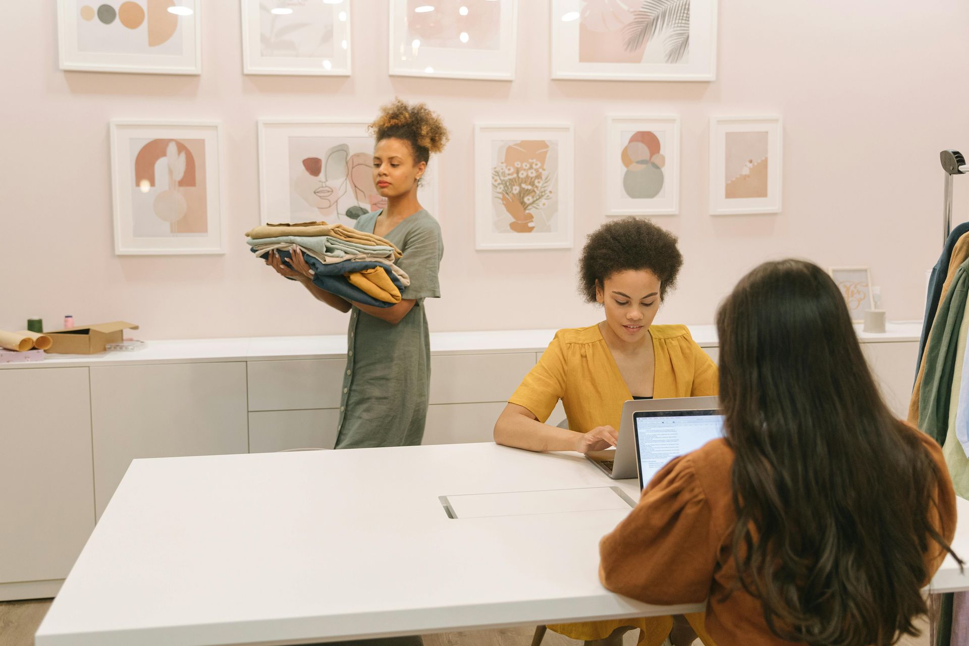 Art studio with three people working at a large white table, surrounded by framed artwork on the walls.