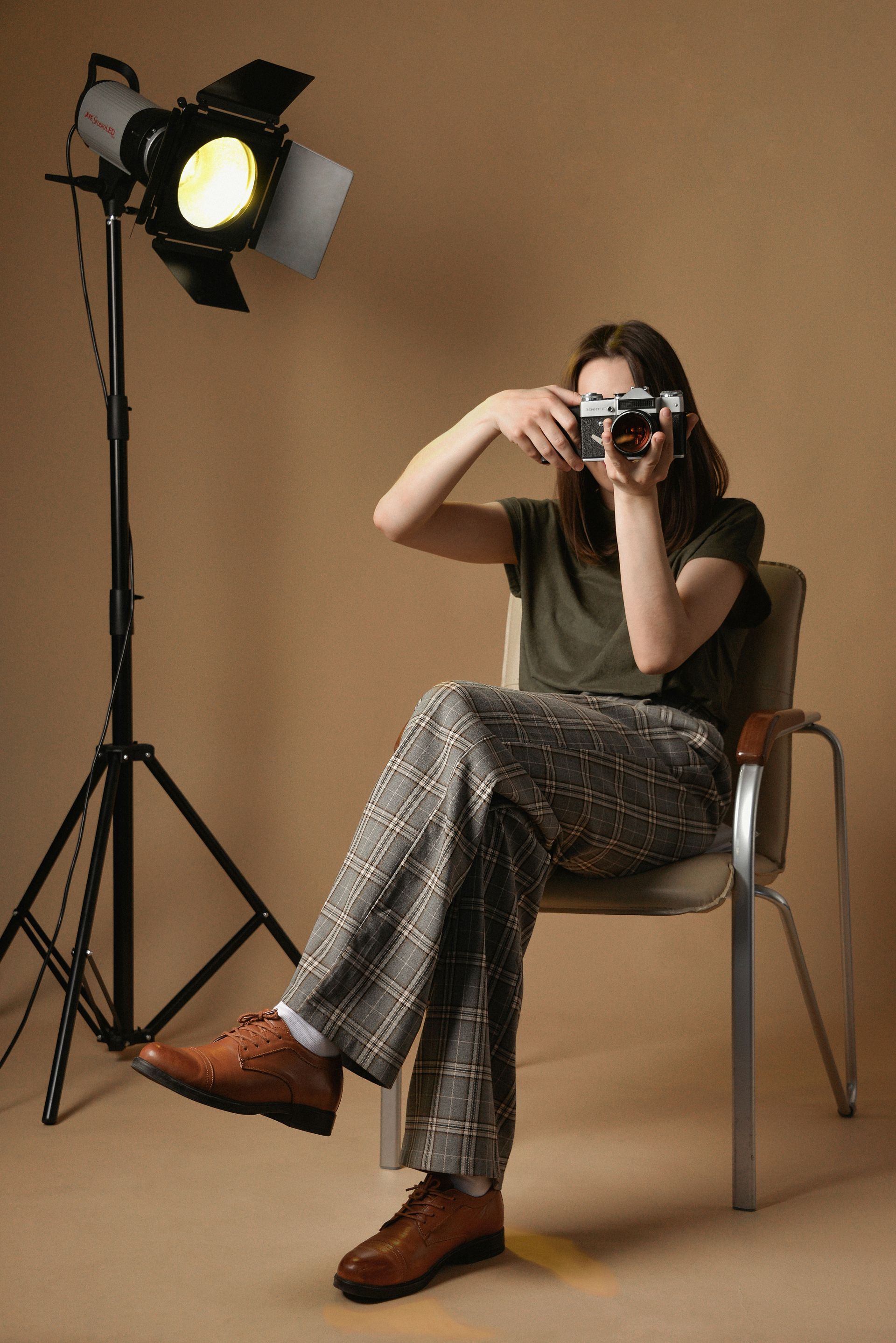 Photographer seated on a chair, holding a camera, beside a lit studio light against a plain backdrop