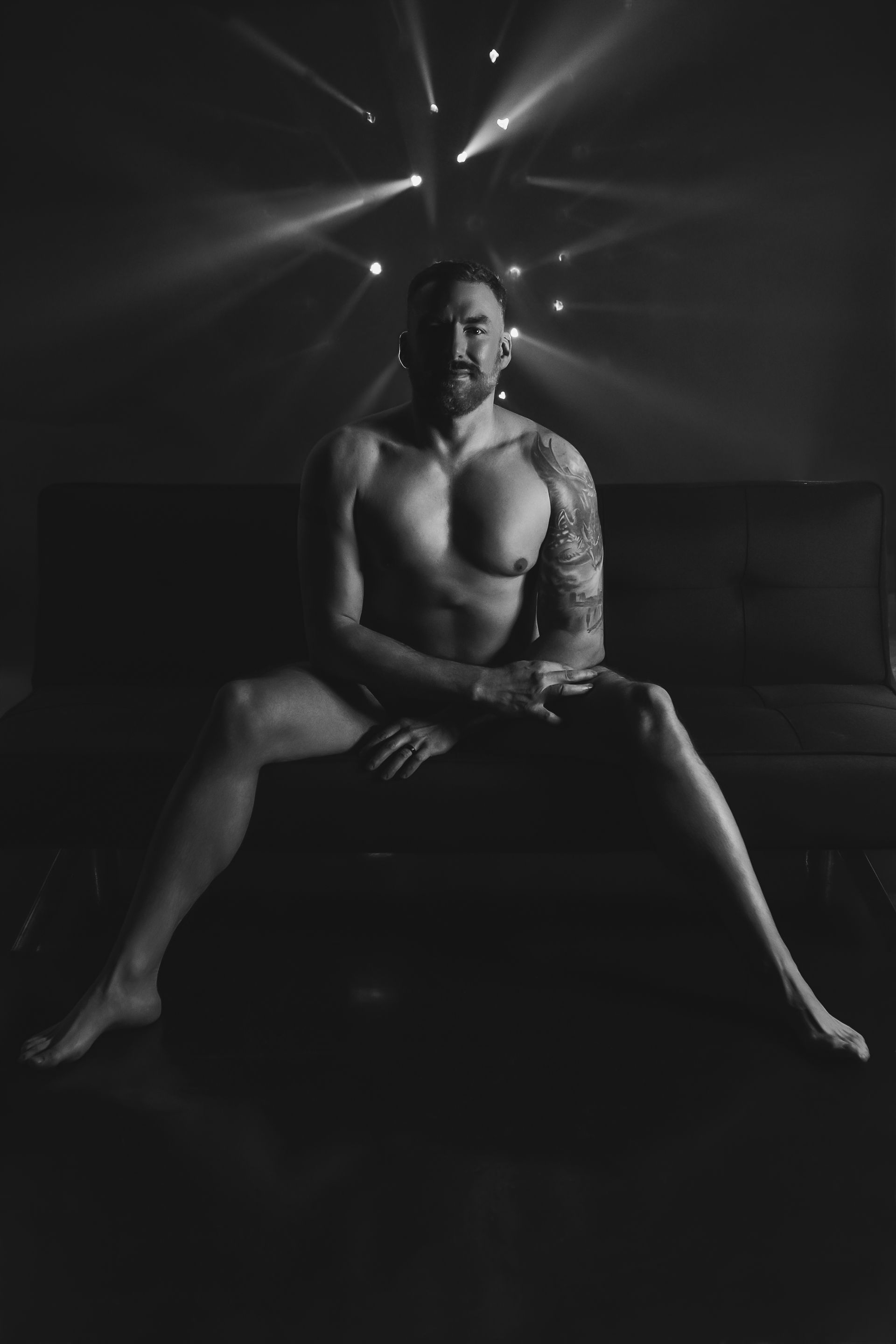 Black-and-white portrait of a shirtless man seated on a couch with legs apart, under dramatic backlighting.