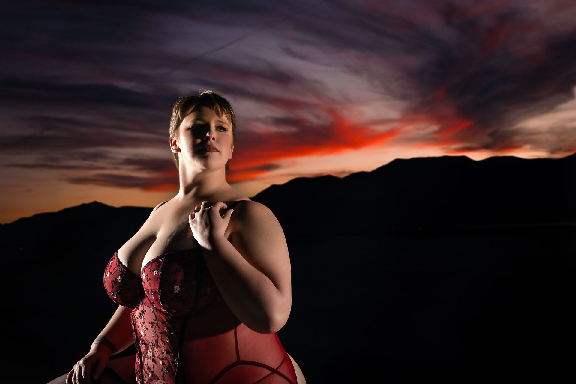 Woman in red lingerie posing against a dramatic sunset sky and mountain silhouettes