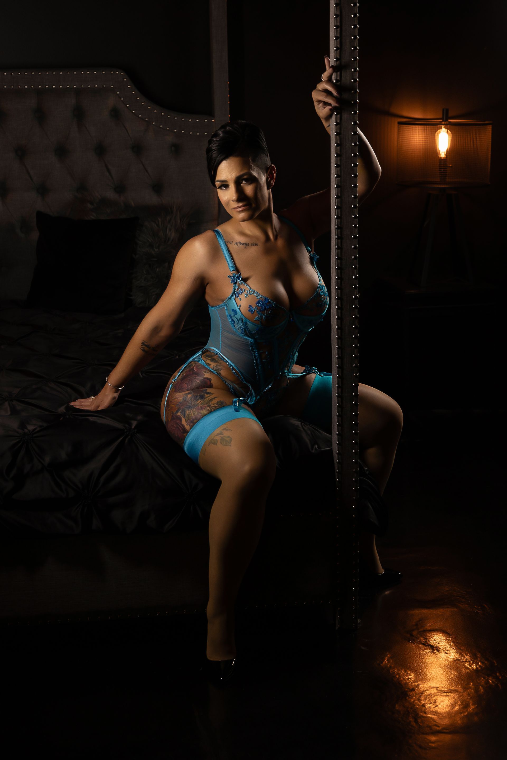 Woman in blue lingerie posing on a pole in a dimly lit room with warm orange light.