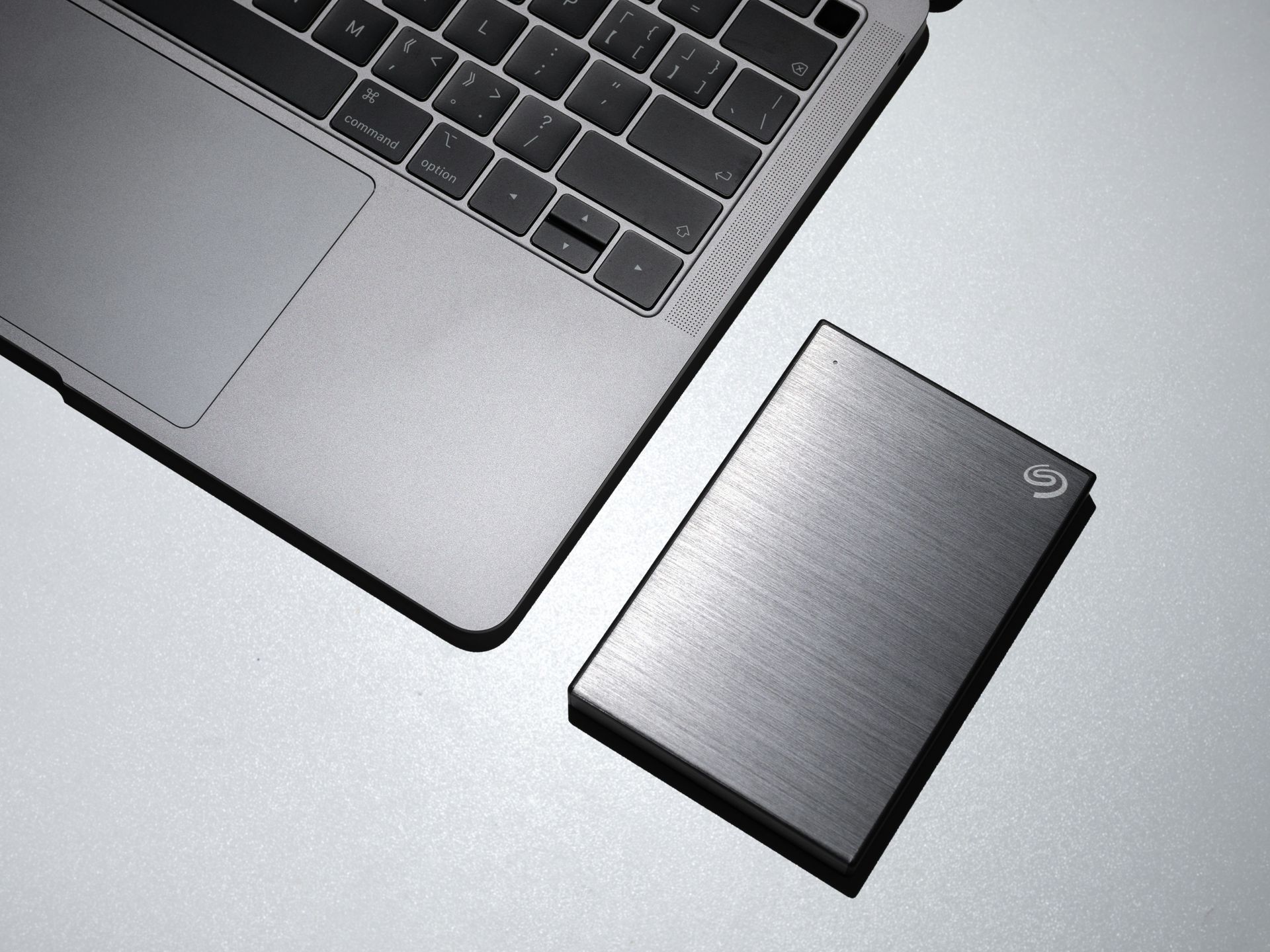 Laptop corner beside a silver external hard drive on a light gray surface