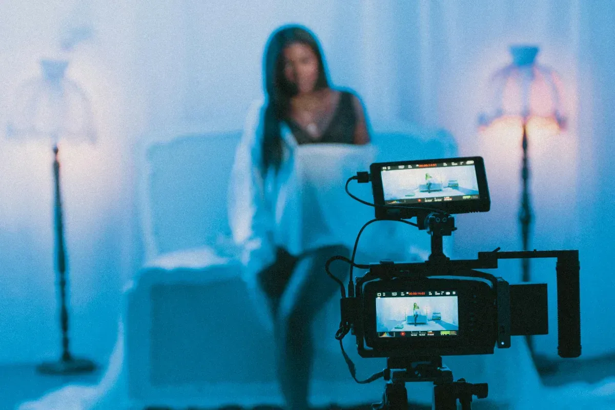Camera filming a woman in a blue-lit studio with two monitors visible in the foreground