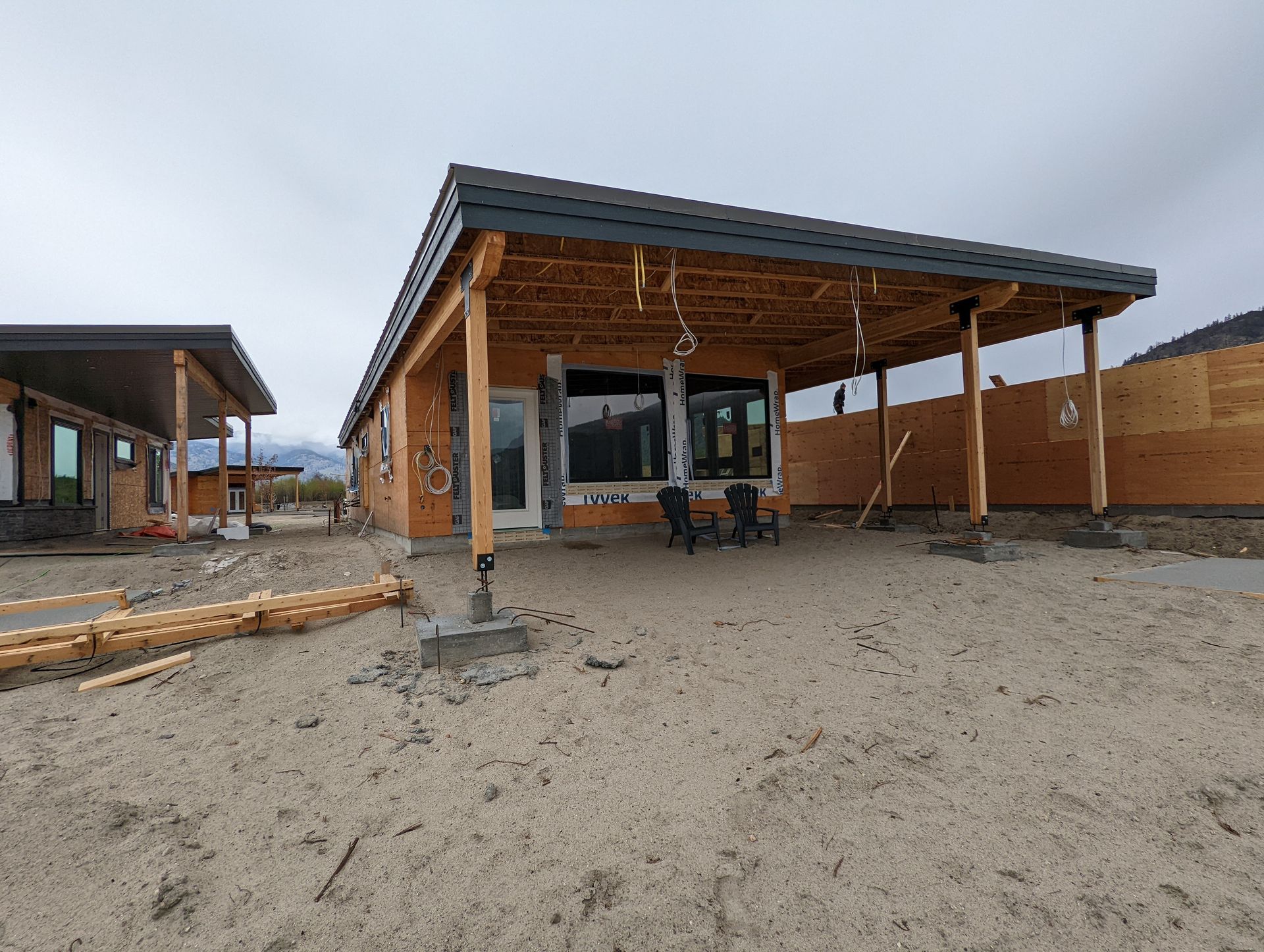 Construction site with wooden buildings featuring covered porches, sandy ground, and mountains in the background under an overcast sky.
