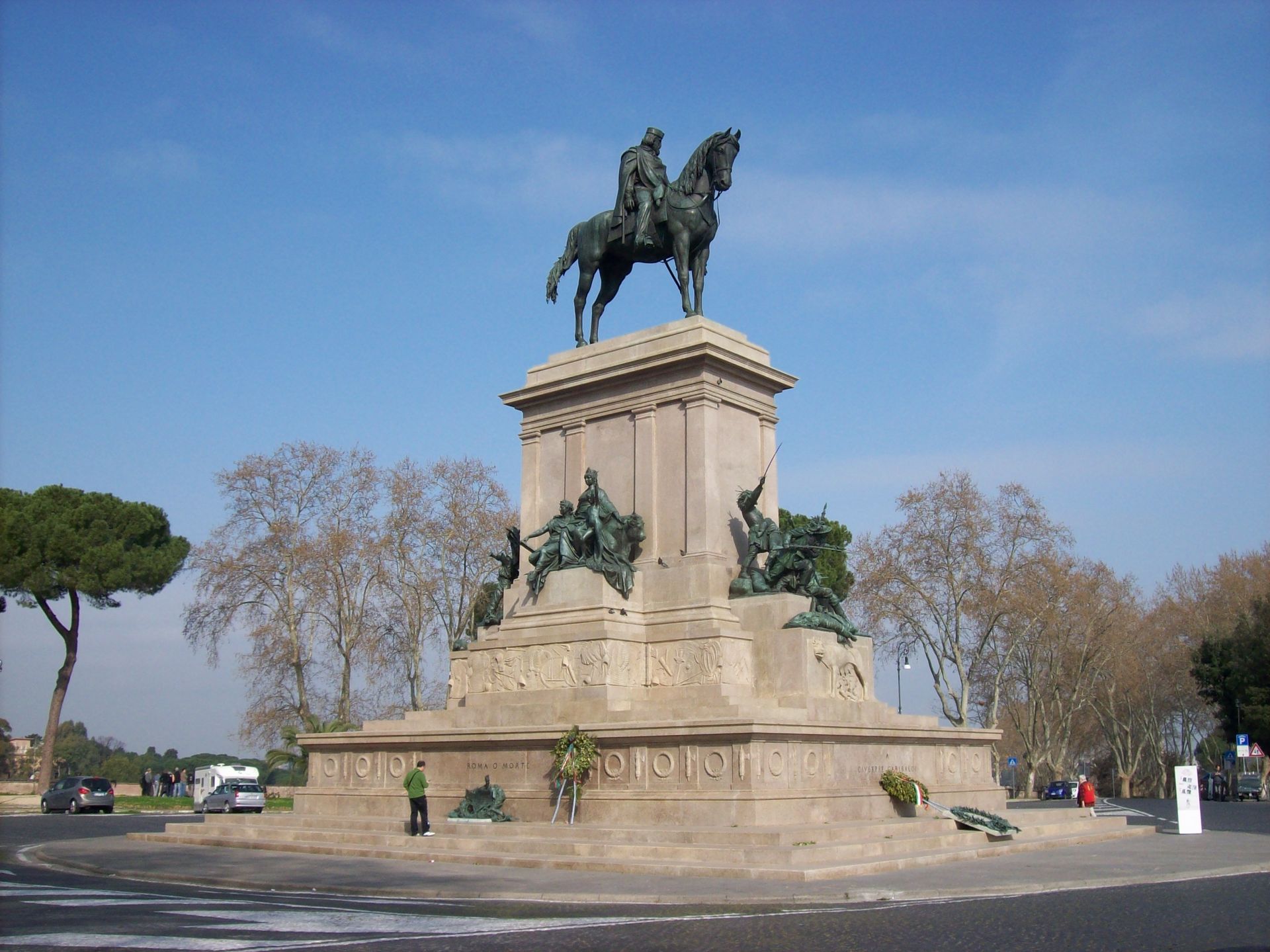 Equestrian statue of a man atop a horse on a large stone base in Rome, Italy. Blue sky, cars, and trees in the background.