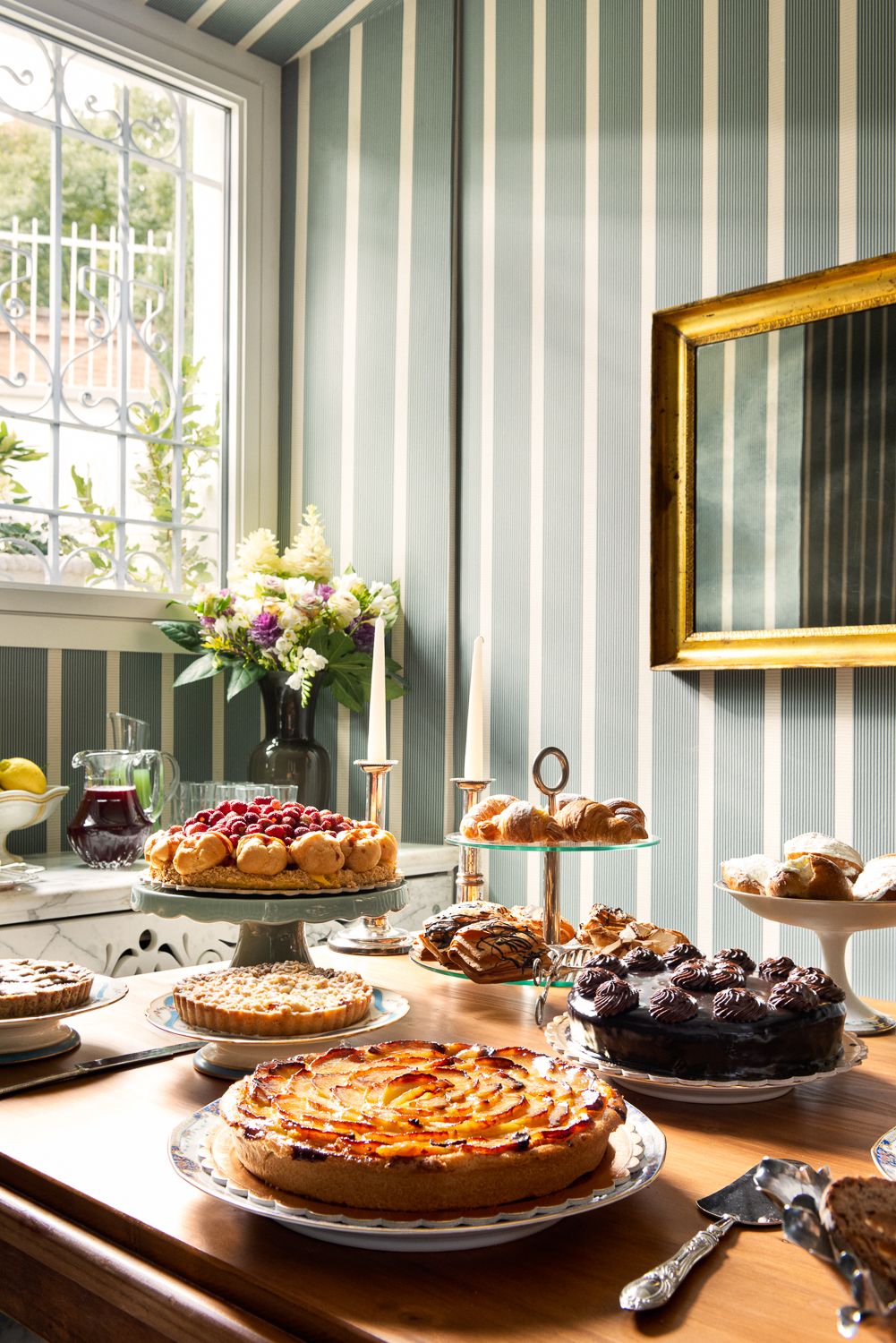 Table laden with cakes and pastries in a room with striped wallpaper and a window.