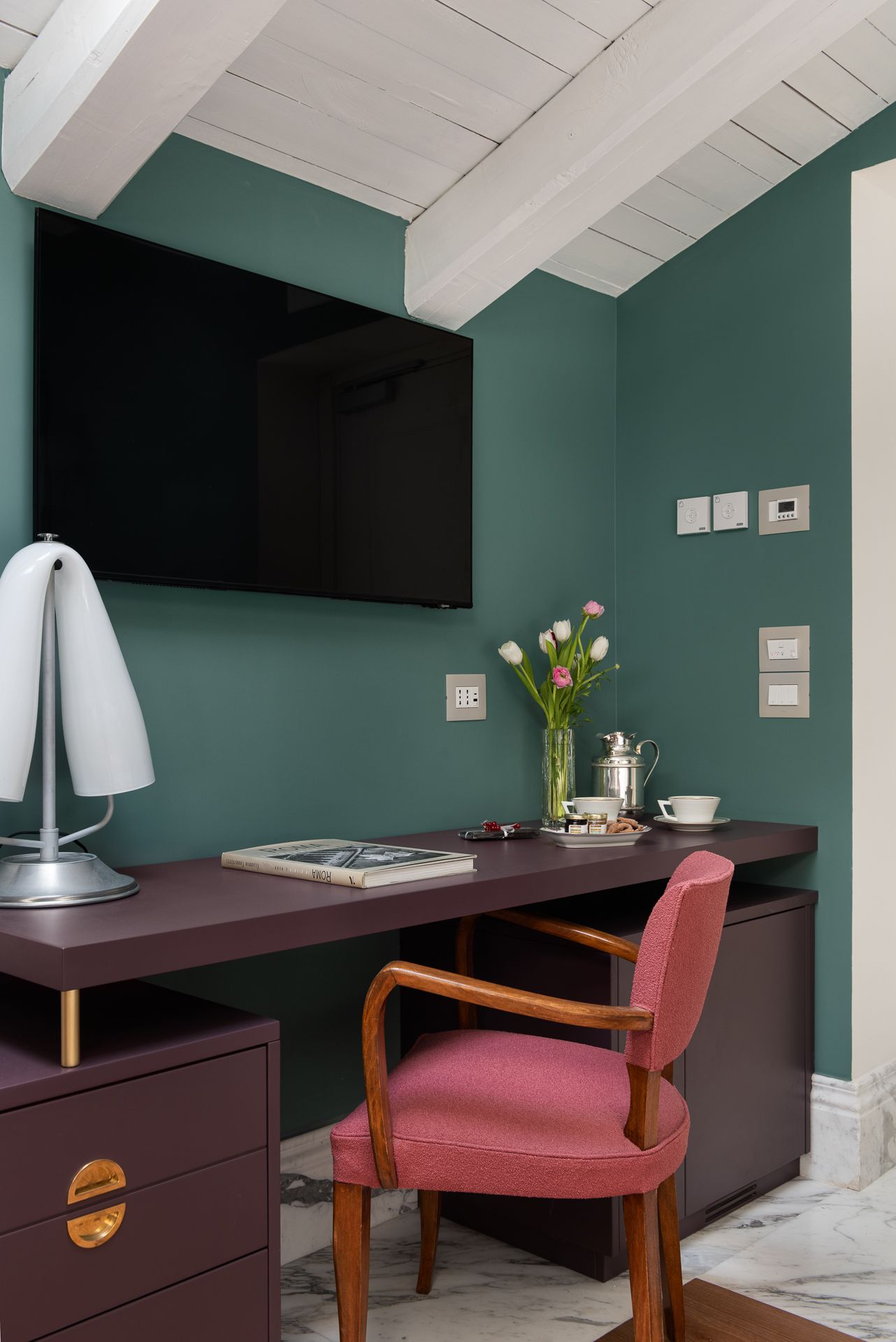 Desk with TV, lamp, and chair in a room with teal walls and white ceiling beams.