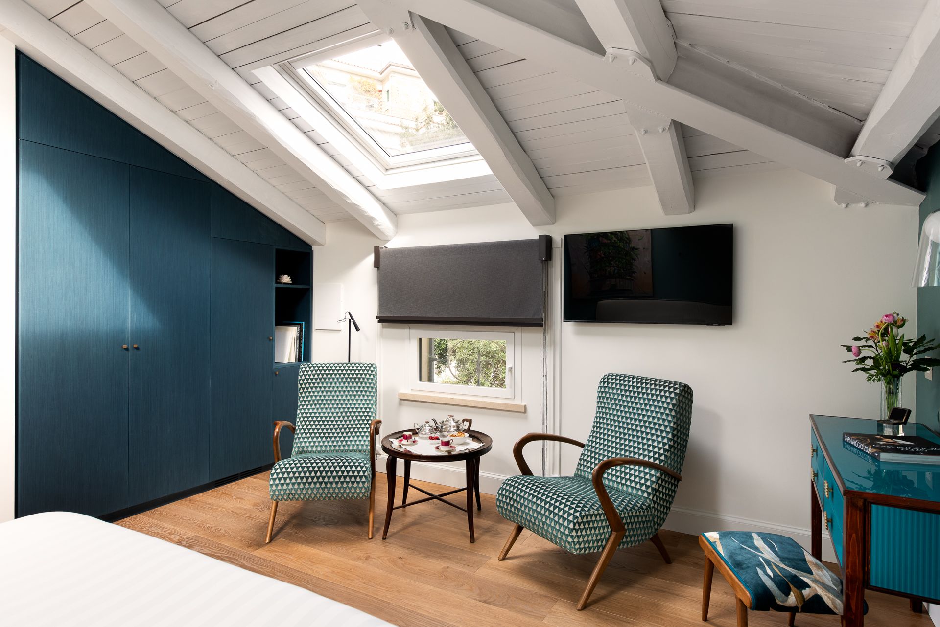 Attic bedroom with teal built-in closet, patterned armchairs, skylight, and small table.