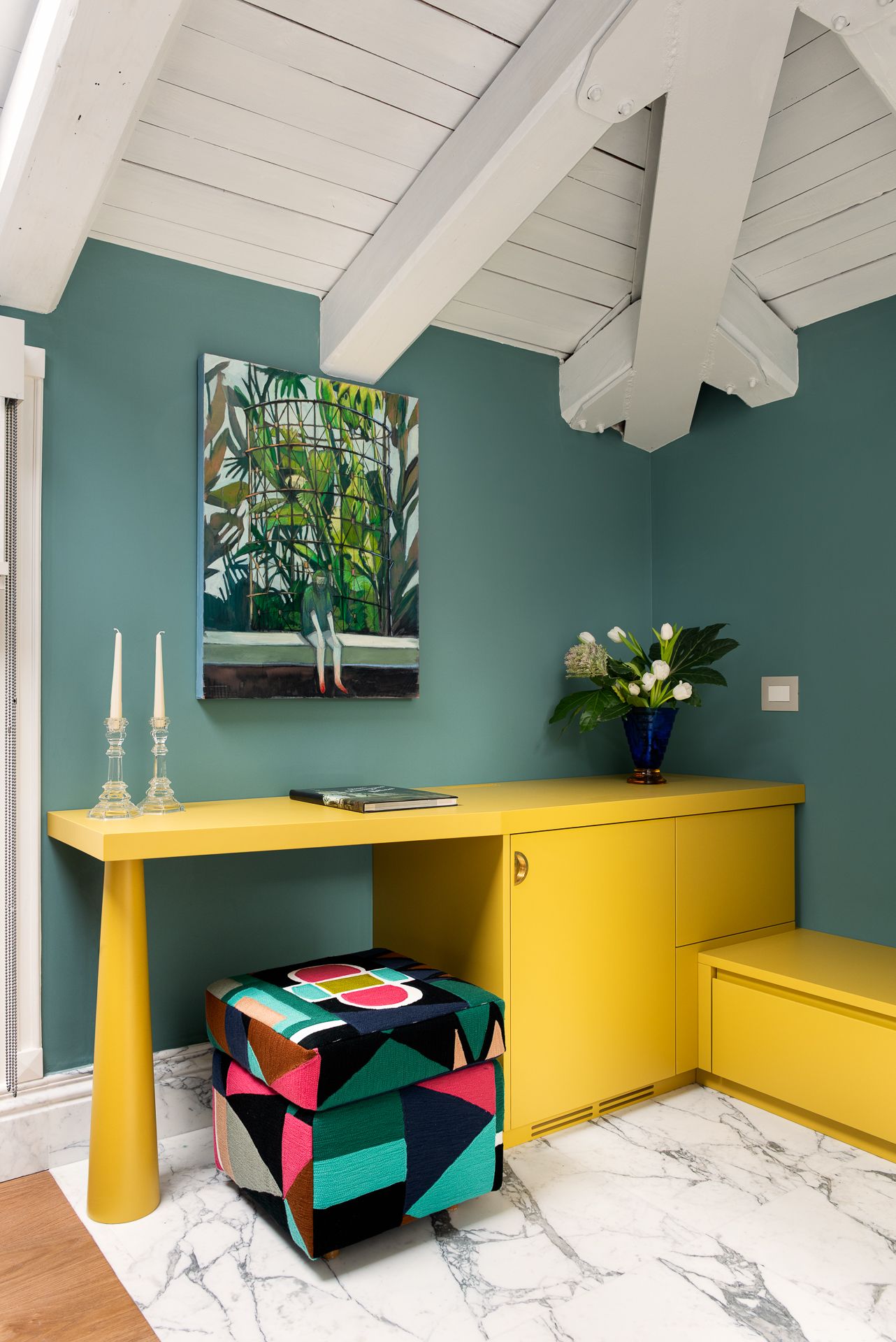 Teal walls, yellow desk, colorful ottoman, artwork, and flowers in a bright room with a white beam ceiling.