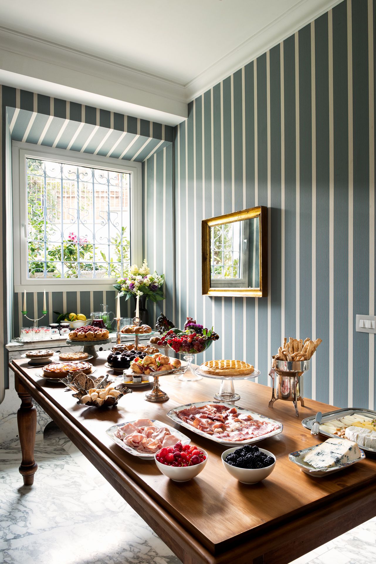 Long wooden table laden with food in a room with striped wallpaper and a window.