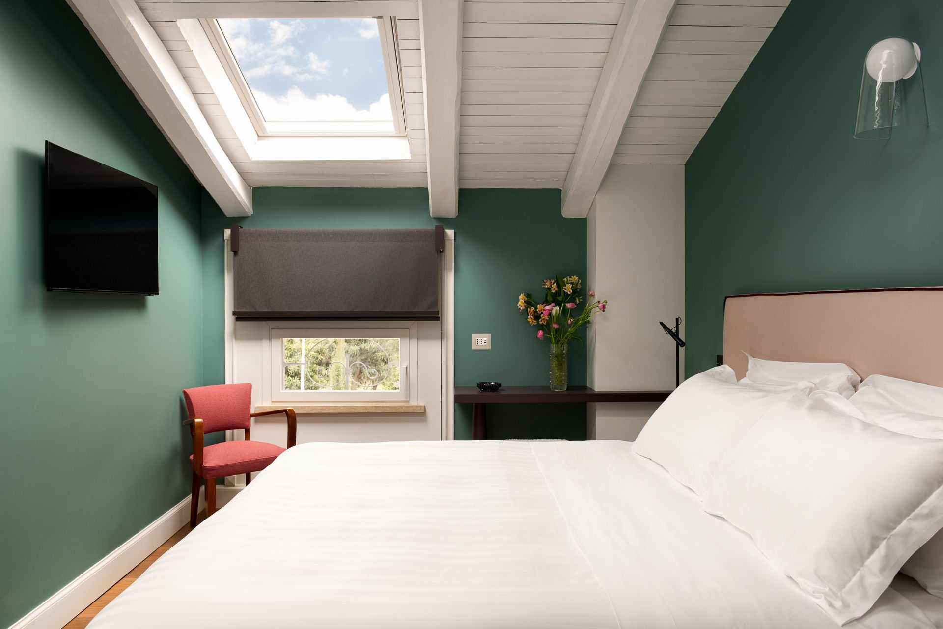 Bedroom with green walls, white bedding, exposed beams, skylight, and small pink chair.
