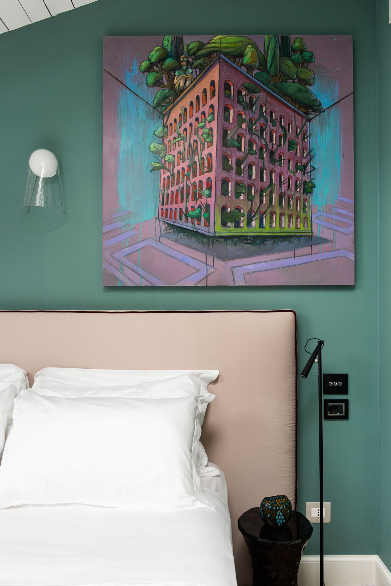 Bedroom with teal walls, pink headboard, white bedding, and artwork of a building covered in greenery.