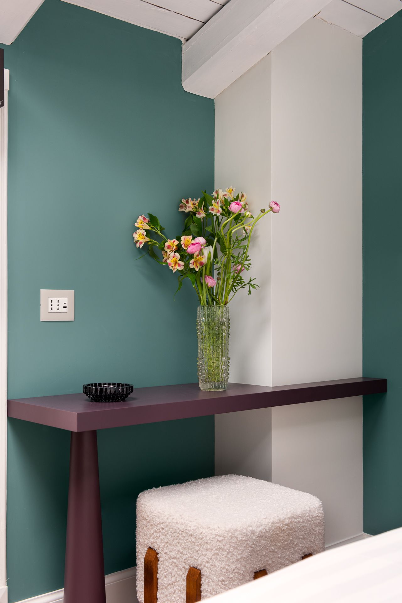 Corner with purple table, vase of flowers, white stool, and teal wall.