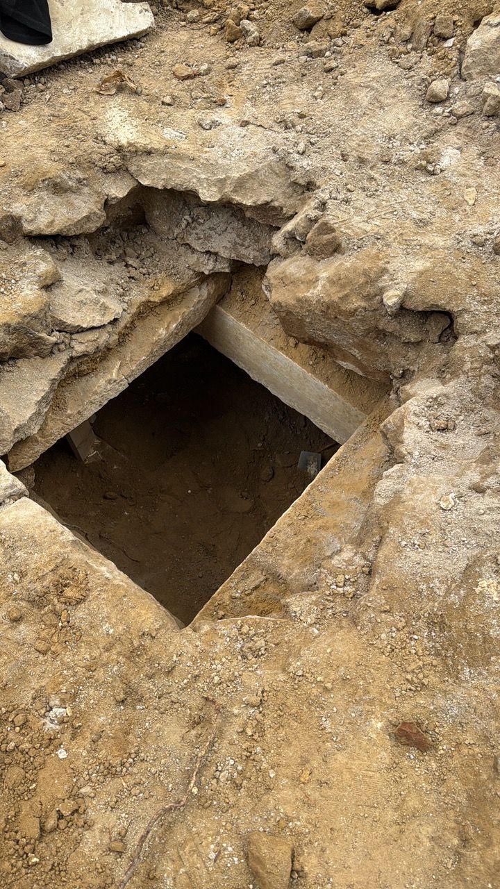 A square hole in the ground reveals a dark interior with a concrete frame. The surrounding soil is tan.