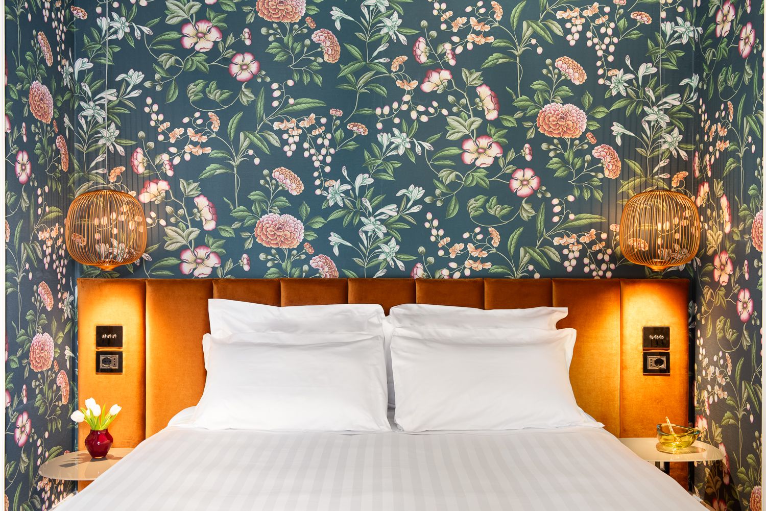Bedroom with floral wallpaper, orange headboard, white bedding, bedside tables with lamps.
