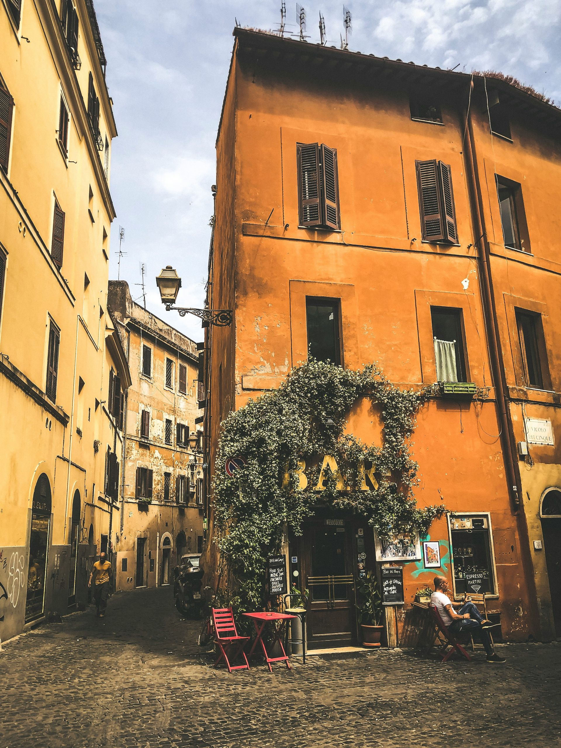 Orange buildings on cobblestone street, entrance to a bar covered in green vines. People at tables.