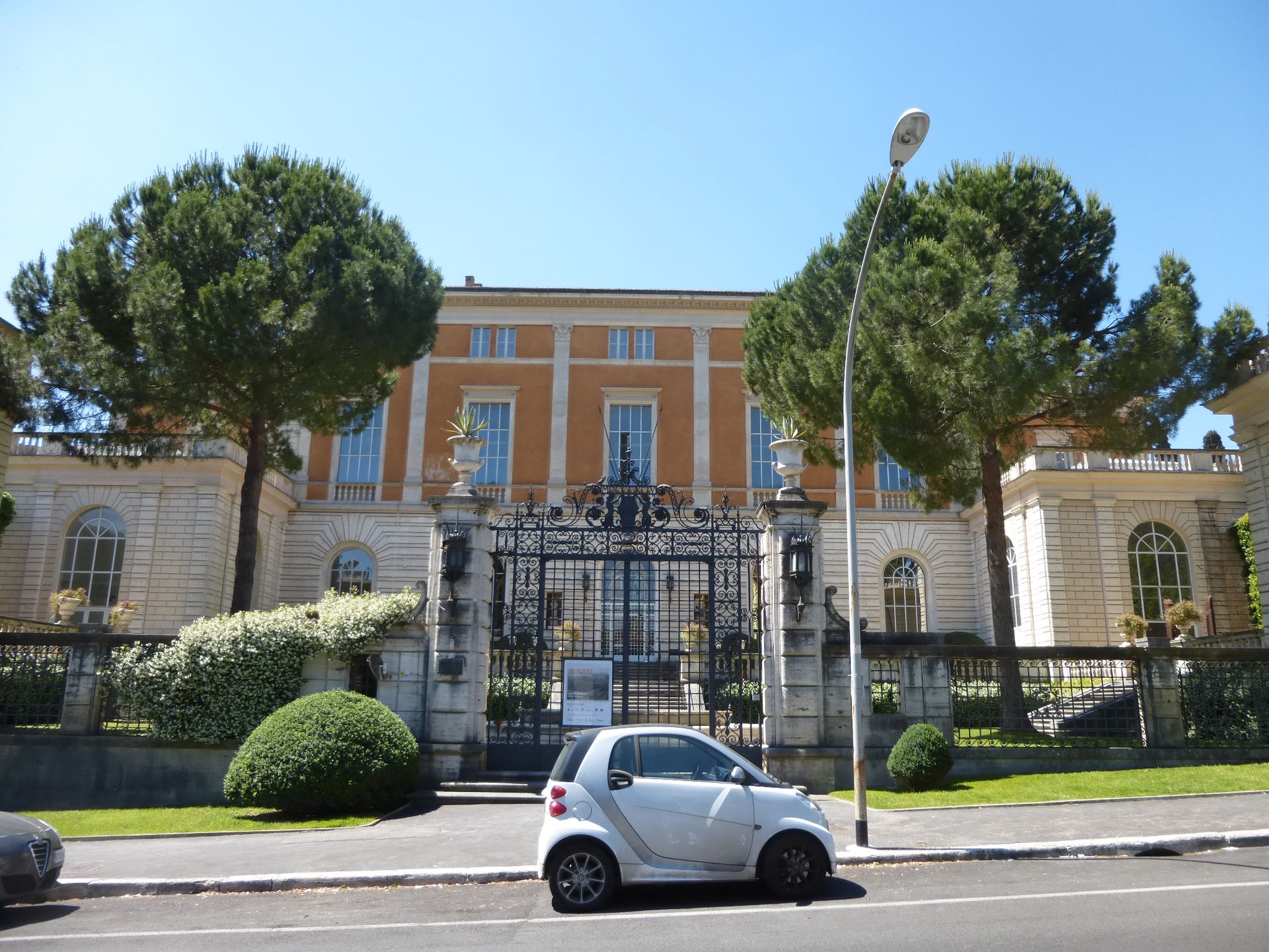 Un edificio arancione chiaro con un cancello in ferro battuto nero, una piccola auto bianca e alberi.
