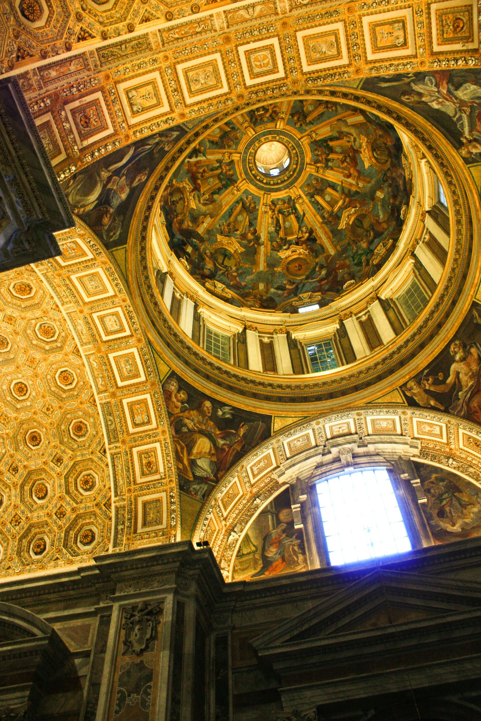 Ornate, golden-hued dome ceiling, decorated with paintings and architectural details. Bright window in lower right.