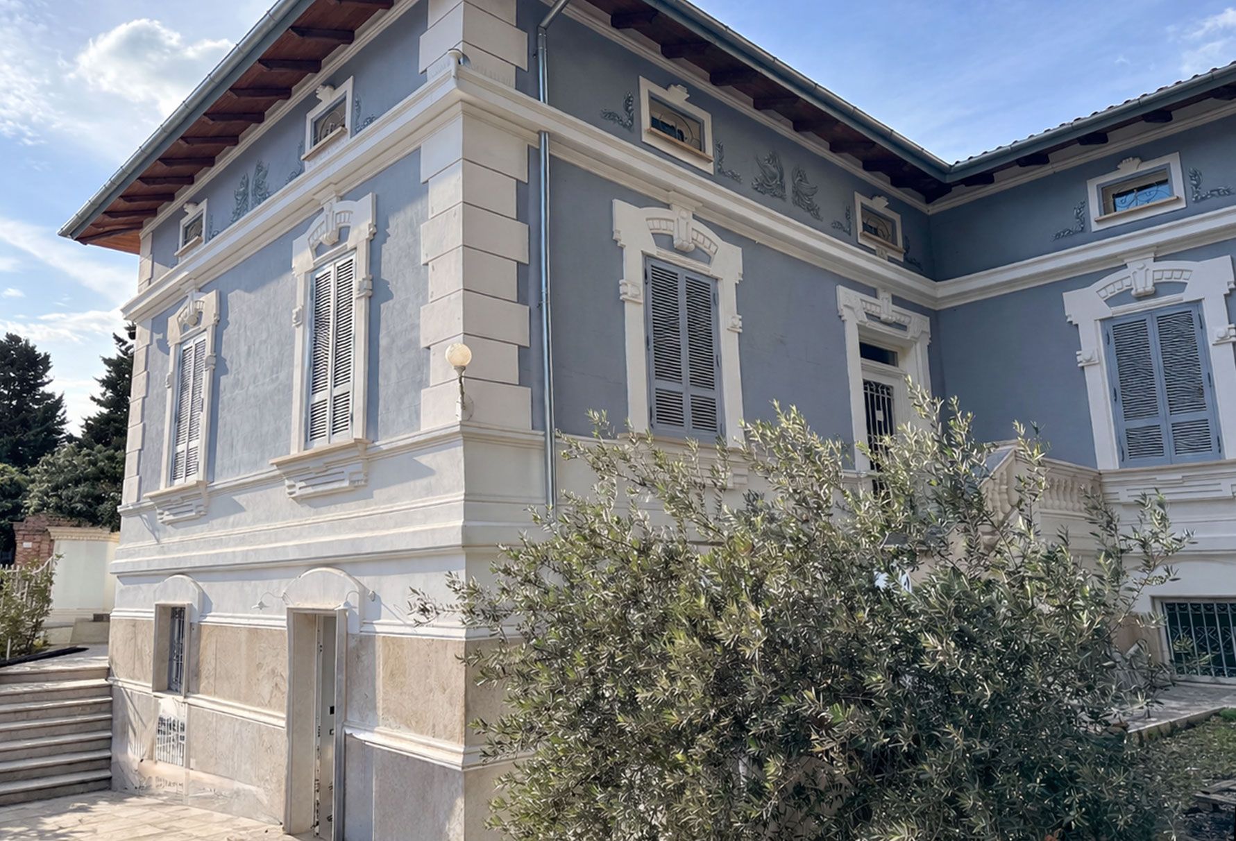 Two-story blue building with white trim, shutters, and brown roof under a partly cloudy sky.