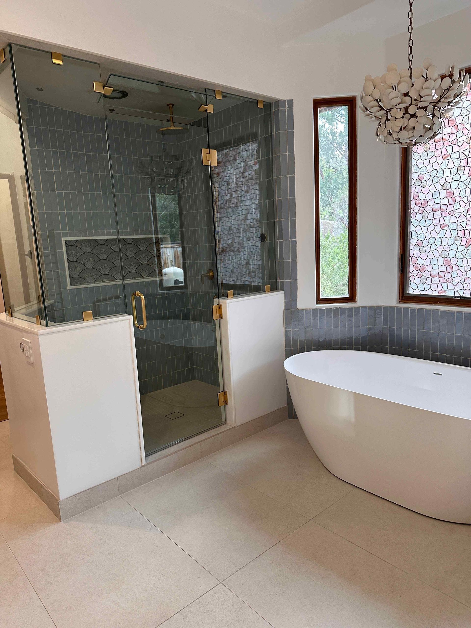 Modern bathroom with glass shower, white freestanding tub, and chandelier by windows