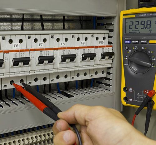 An electrician using a digital multimeter to measure voltage on a row of circuit breakers in an electrical panel.