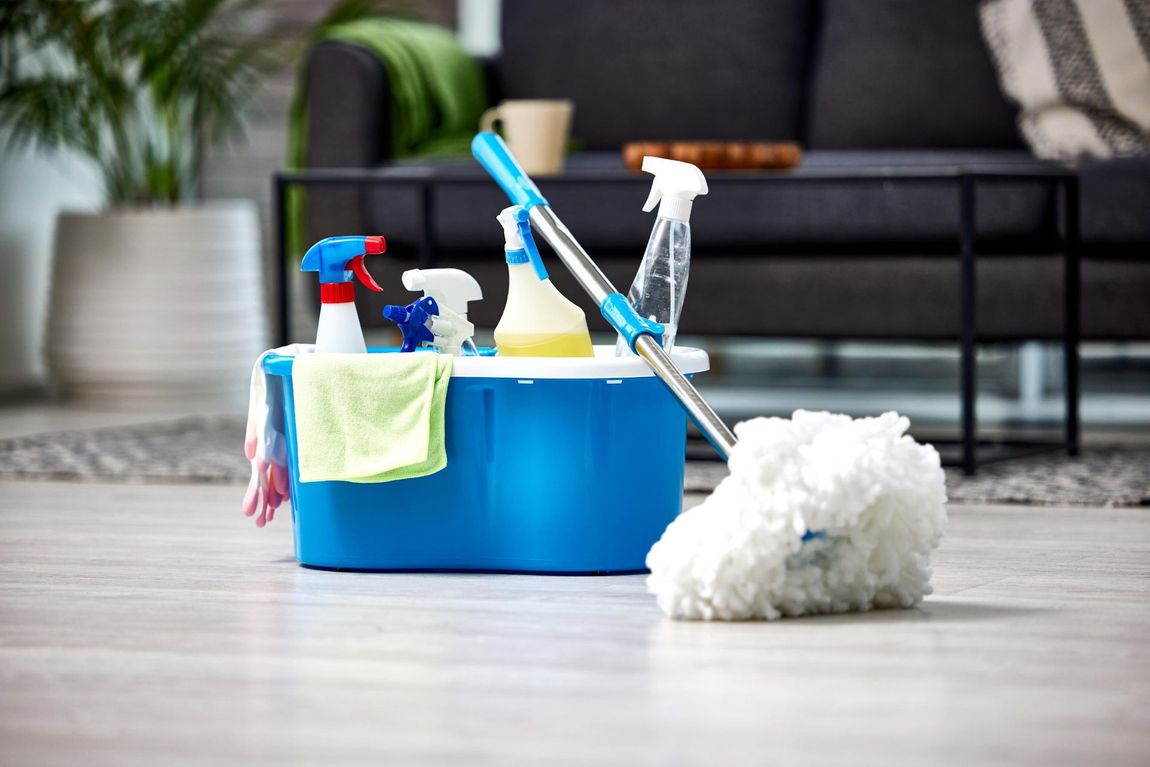 Two cleaners in blue uniforms work in a room with a blue sofa; one mops the wood floor while the other dusts a window.