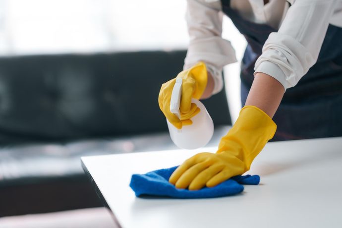 Person wearing yellow cleaning gloves wipes a white surface with a blue cloth while holding a spray bottle.