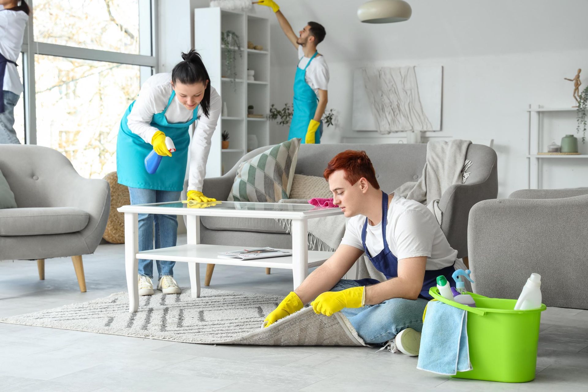 A diverse group of cleaners in teal aprons and yellow gloves tidy a bright, modern living room.