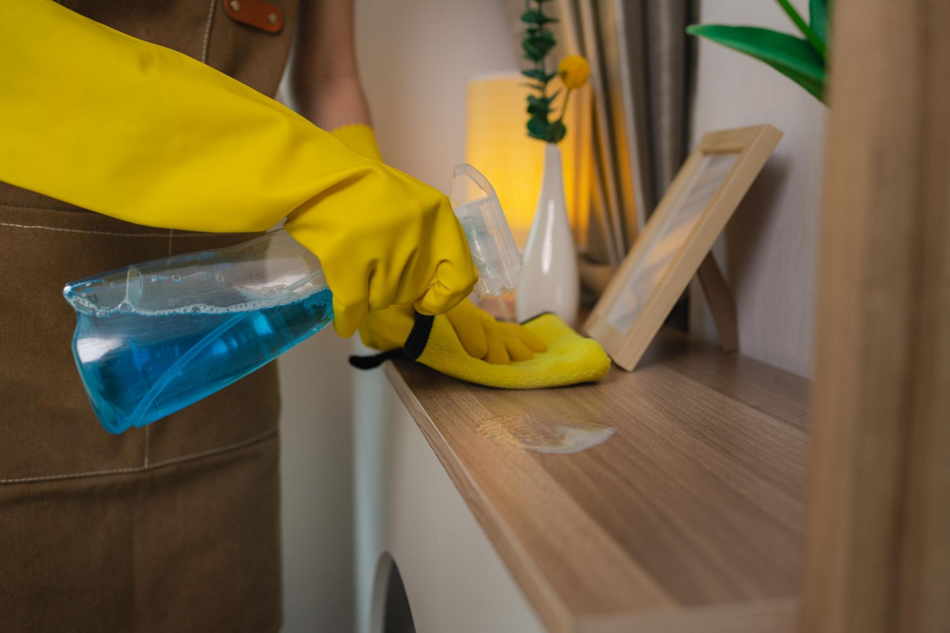 A person wearing yellow rubber gloves wipes a wooden shelf with a cleaning spray and cloth.