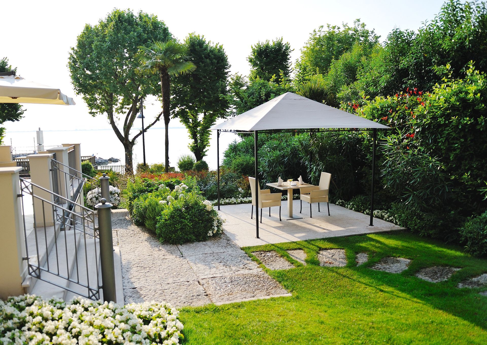 Patio con tavolo e sedie sotto una tettoia bianca, circondato da alberi verdi e con vista sull'acqua.