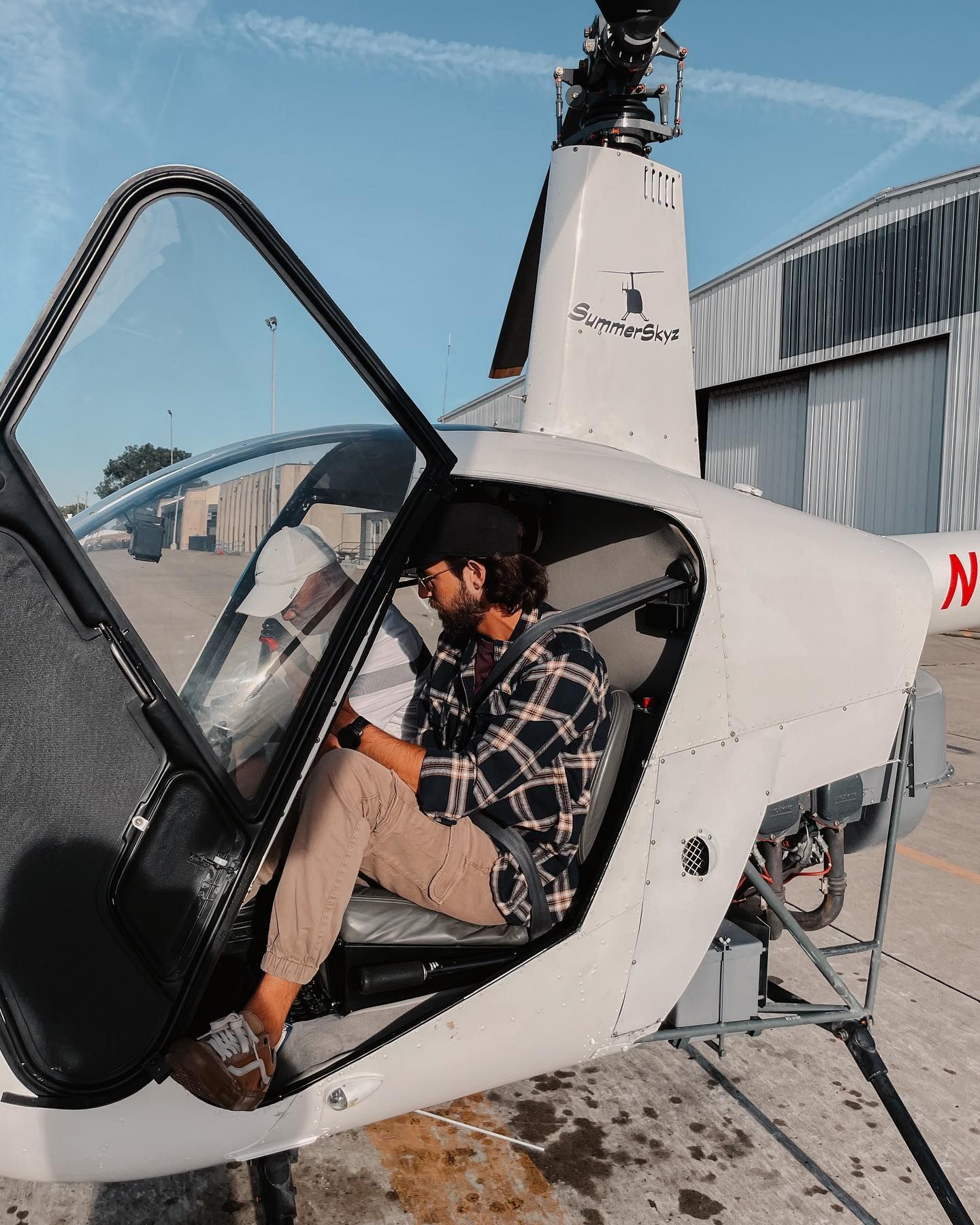 Man in a helicopter cockpit, wearing a plaid shirt and tan pants, door open, sunny day.