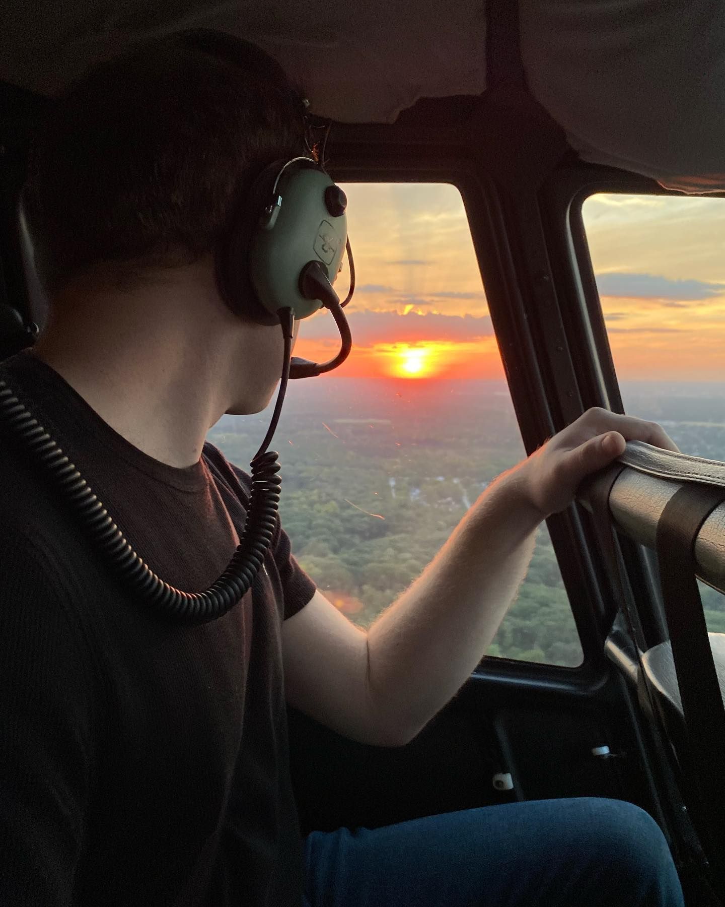 Person in helicopter cockpit watches sunset over trees.